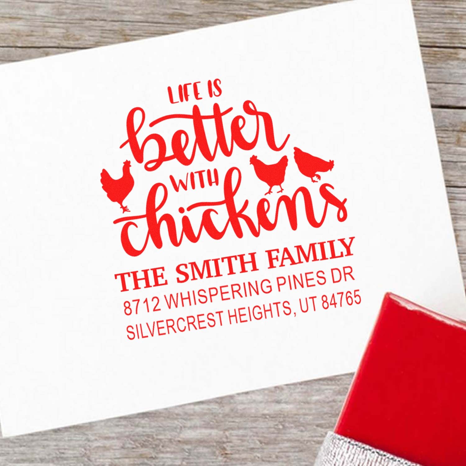 Custom red chicken family address stamp reading Life is better with chickens on white paper beside a red ink pad on rustic wood.