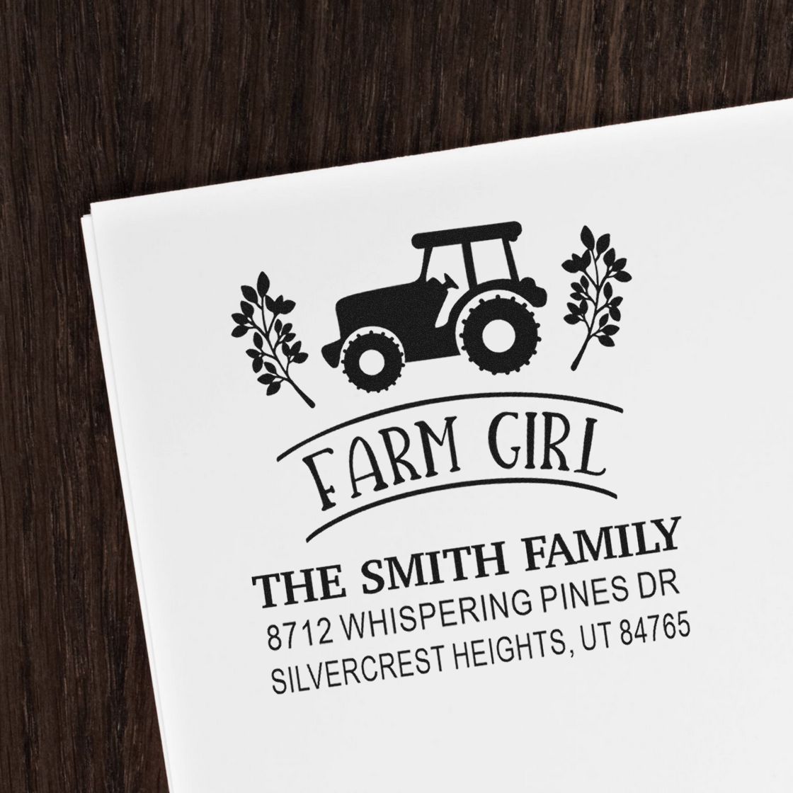 Custom farm girl return address stamp with tractor and leaves on white paper, reading The Smith Family, on a dark wood table.