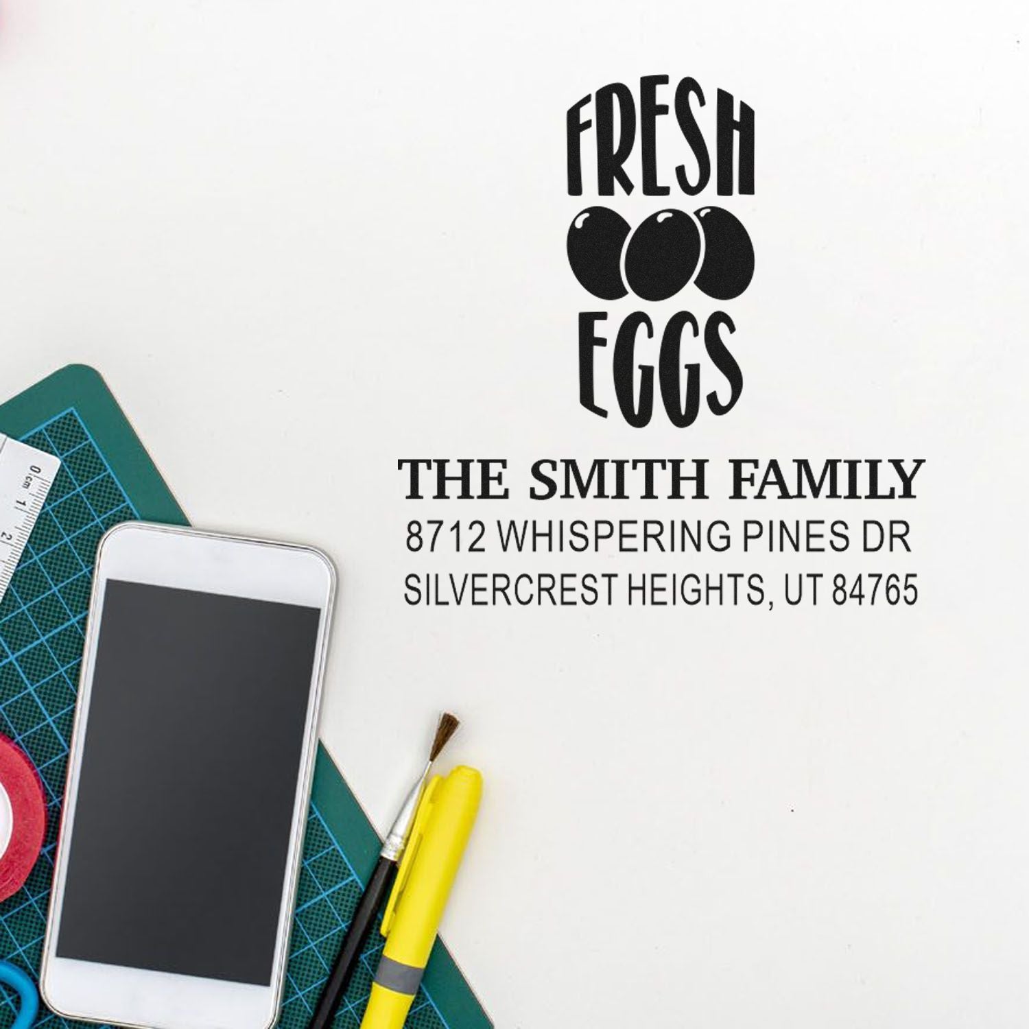 Personalized return address stamp design with Fresh Eggs and three eggs for The Smith Family, displayed on a craft desk with cutting mat, phone, and tools.