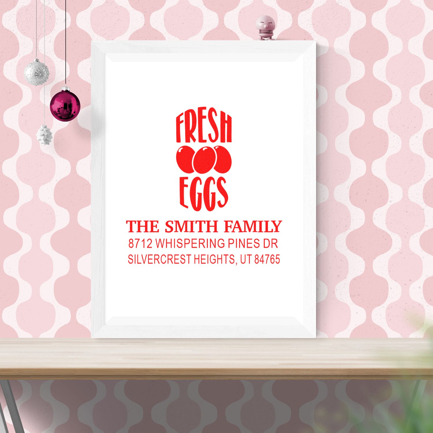Custom holiday stamp reading Fresh Eggs for The Smith Family, framed on a pink patterned wall with hanging ornaments.