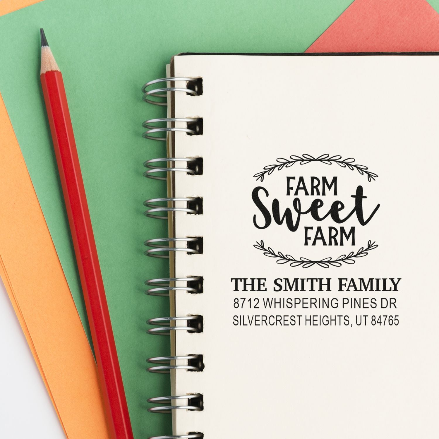 Custom Farm Sweet Farm address stamp on a spiral notebook, with a red pencil and colorful paper background.