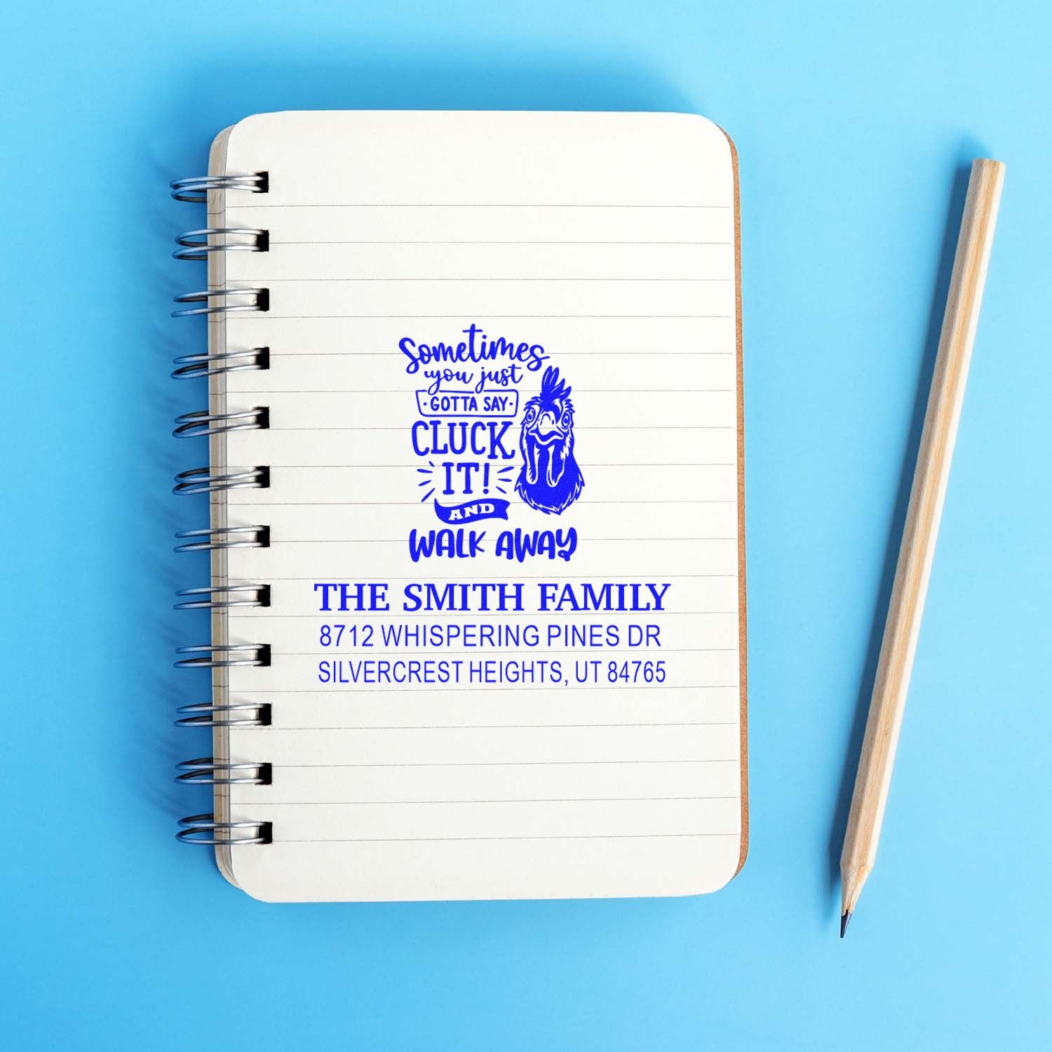 Custom blue self-inking stamp impression on a spiral notebook, featuring a chicken quote and The Smith Family address, with a pencil on a blue background.