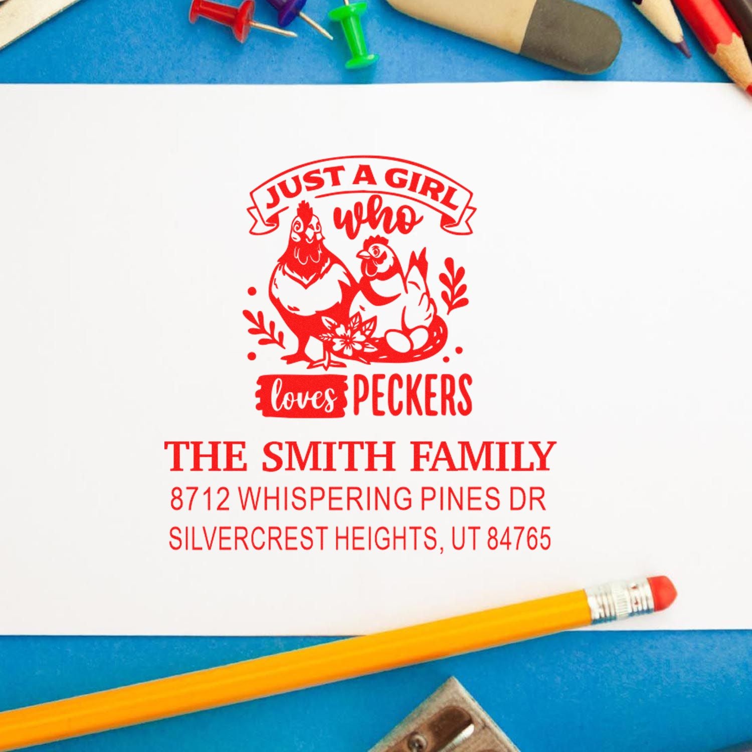 Custom red rubber stamp with chicken graphic and family address on white paper, framed by pencils and push pins on a blue tabletop.