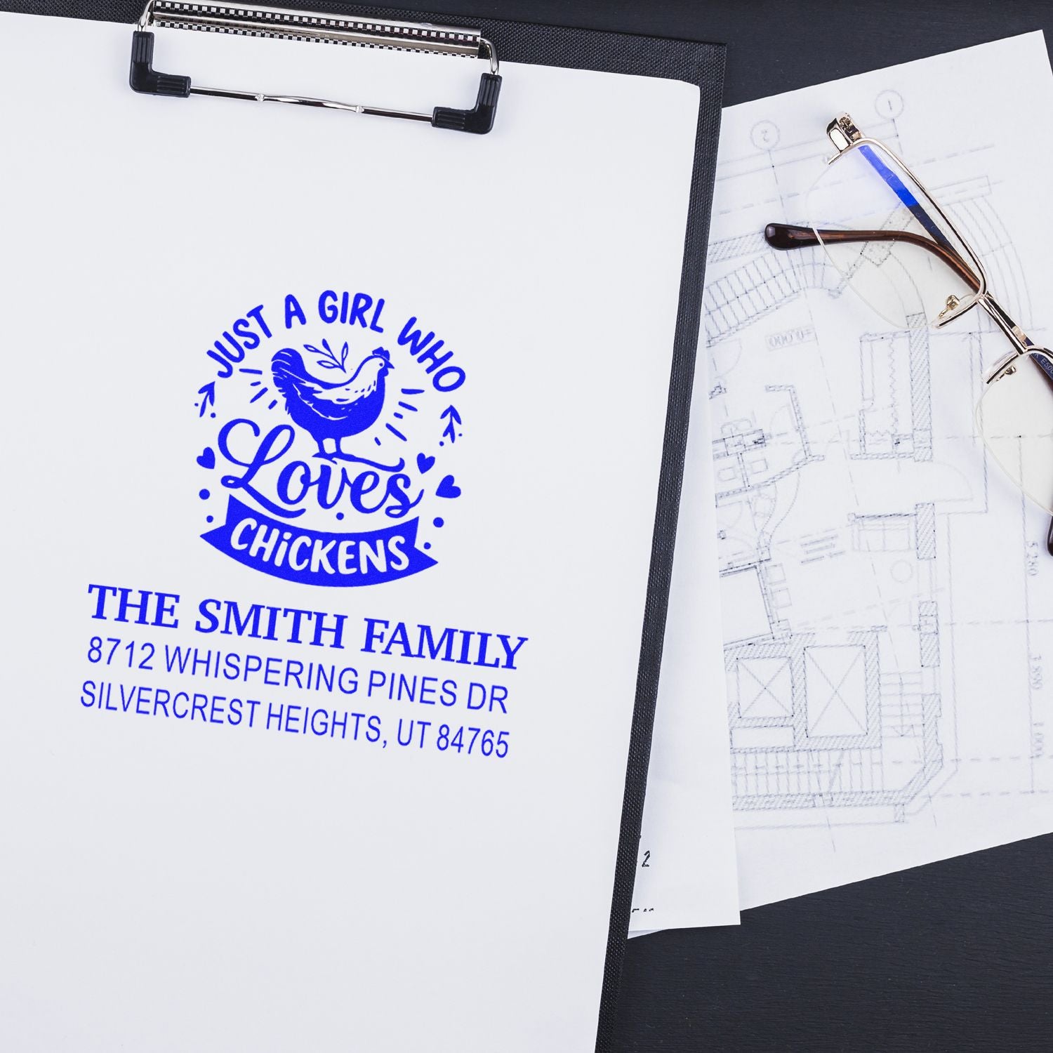 Custom address stamp with chicken design for The Smith Family on clipboard beside eyeglasses and house plans.