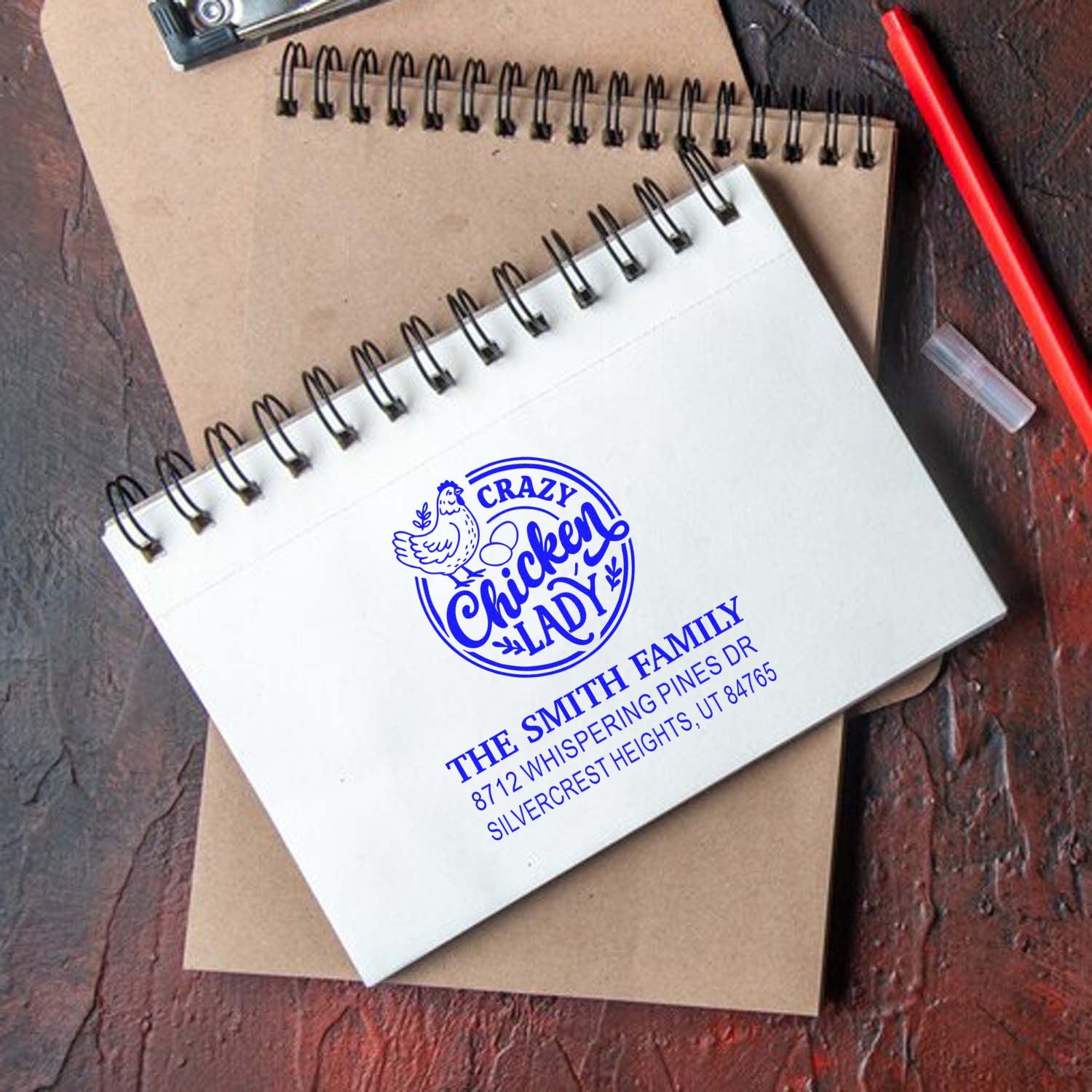 Custom chicken lady address stamp on white spiral notebook, beside kraft notebook and red pen on rustic wooden desk.