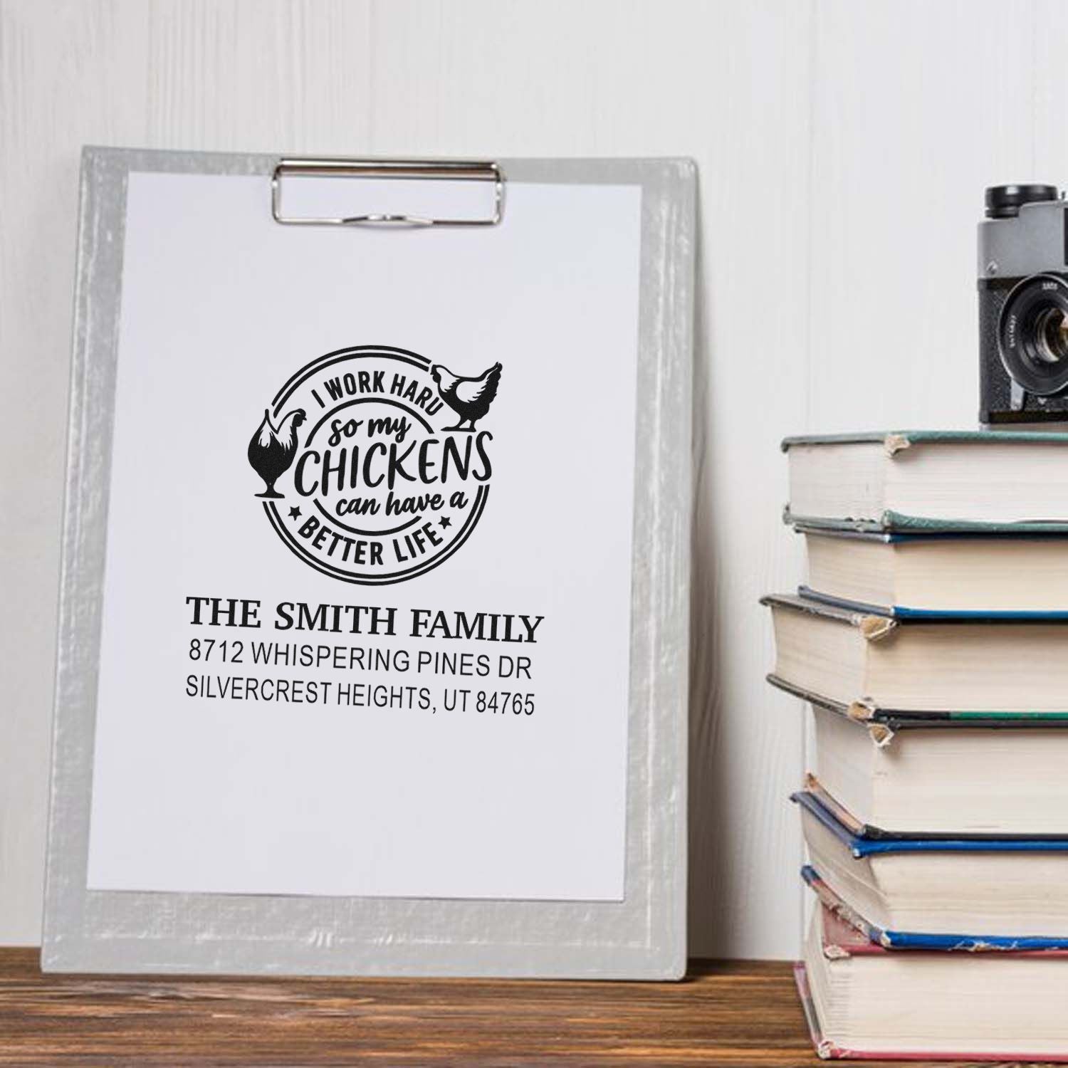 Personalized chicken-themed address stamp impression on clipboard, reading The Smith Family, beside stacked books and a vintage camera.