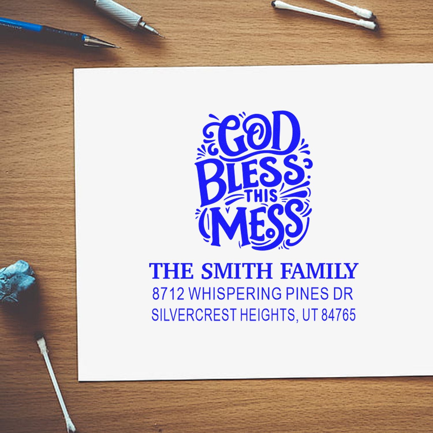 Custom return address stamp reading God Bless This Mess for The Smith Family, displayed on white paper on a wooden desk with pens and cotton swabs.