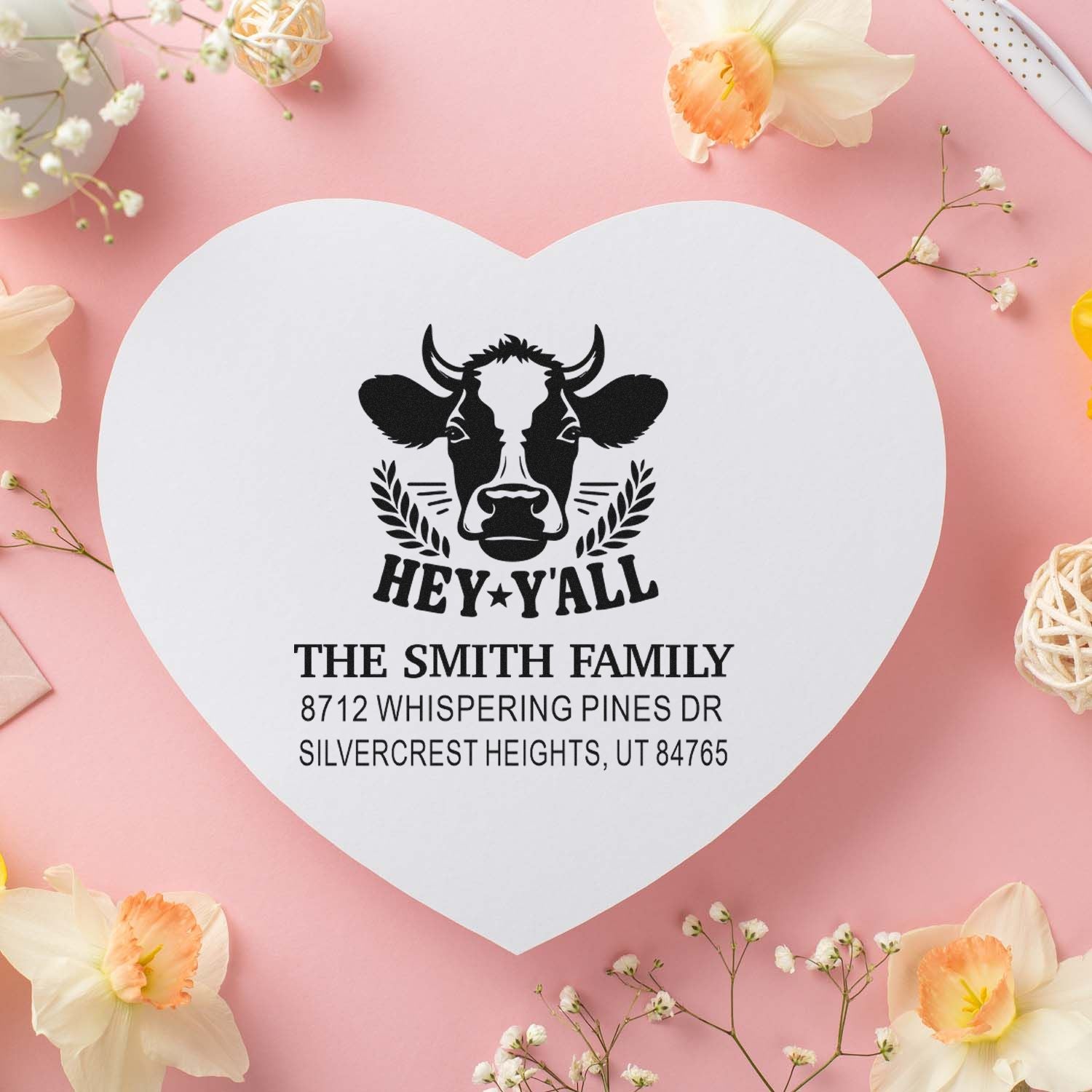 Heart-shaped custom family address stamp featuring a cow graphic, Hey Yall text, and Smith family address on a pink floral flat lay.