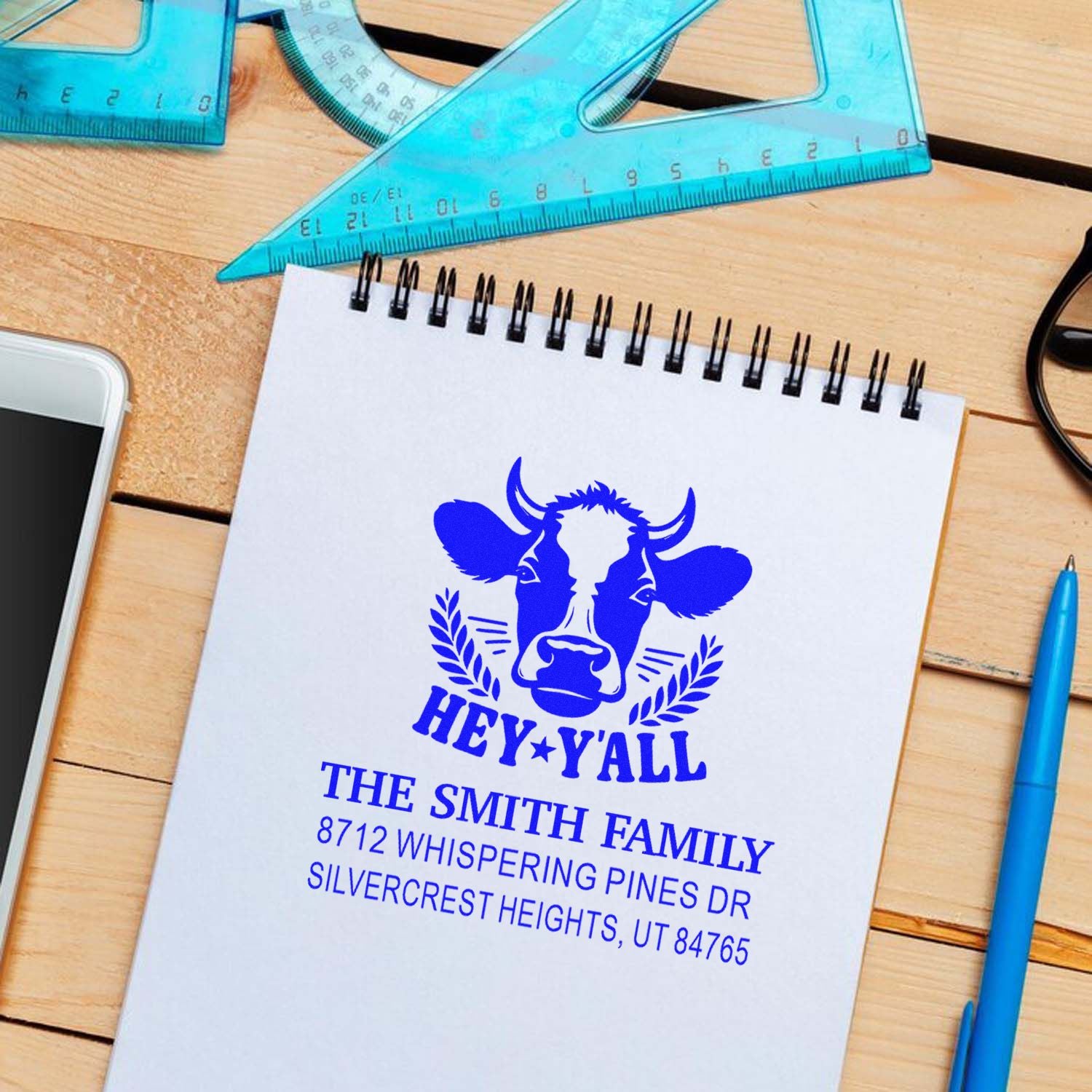 Personalized cow address stamp impression on a spiral notebook, with 'Hey Y'all' and The Smith Family return address on a wooden desk.
