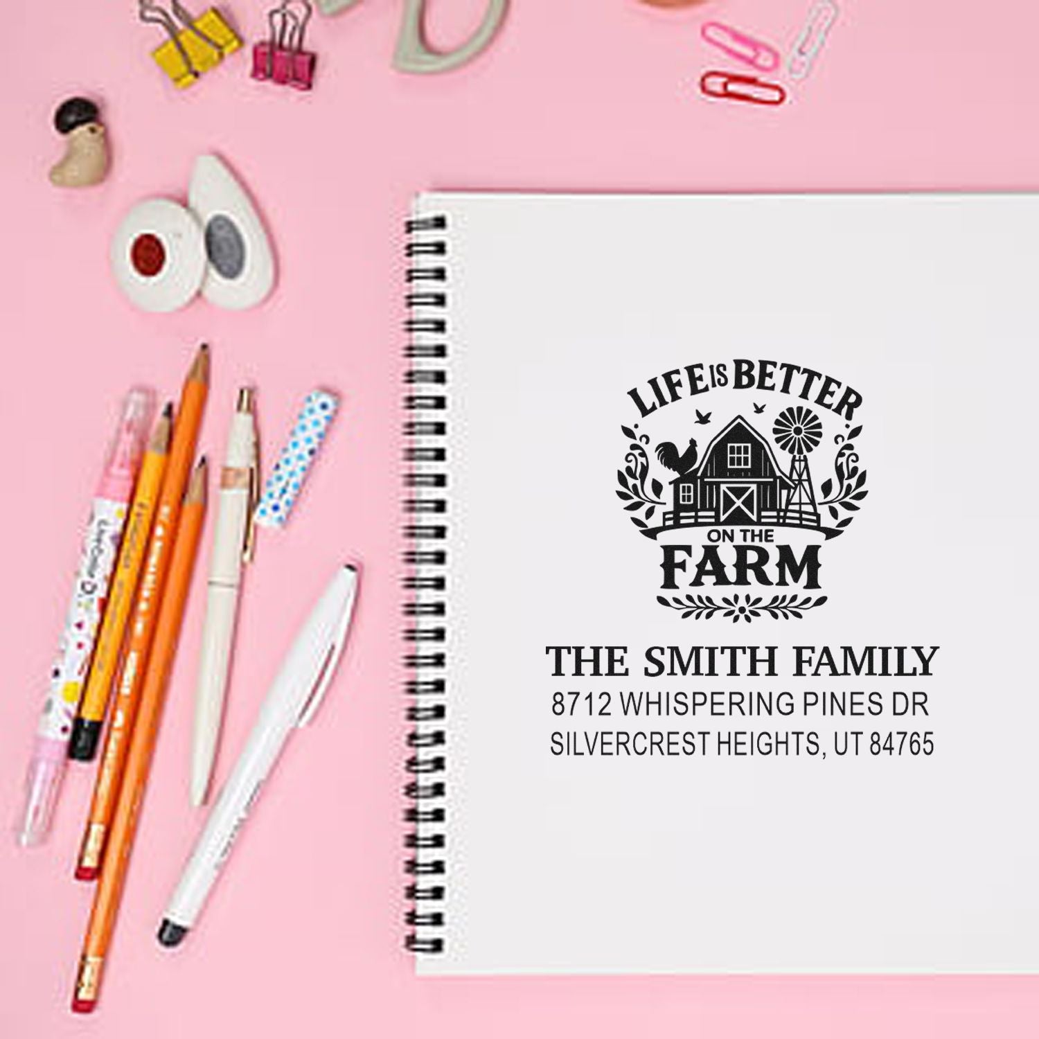 Personalized farm address stamp on a spiral notebook with pens and stationery on a pink desk, featuring Life is Better on the Farm and The Smith Family address.