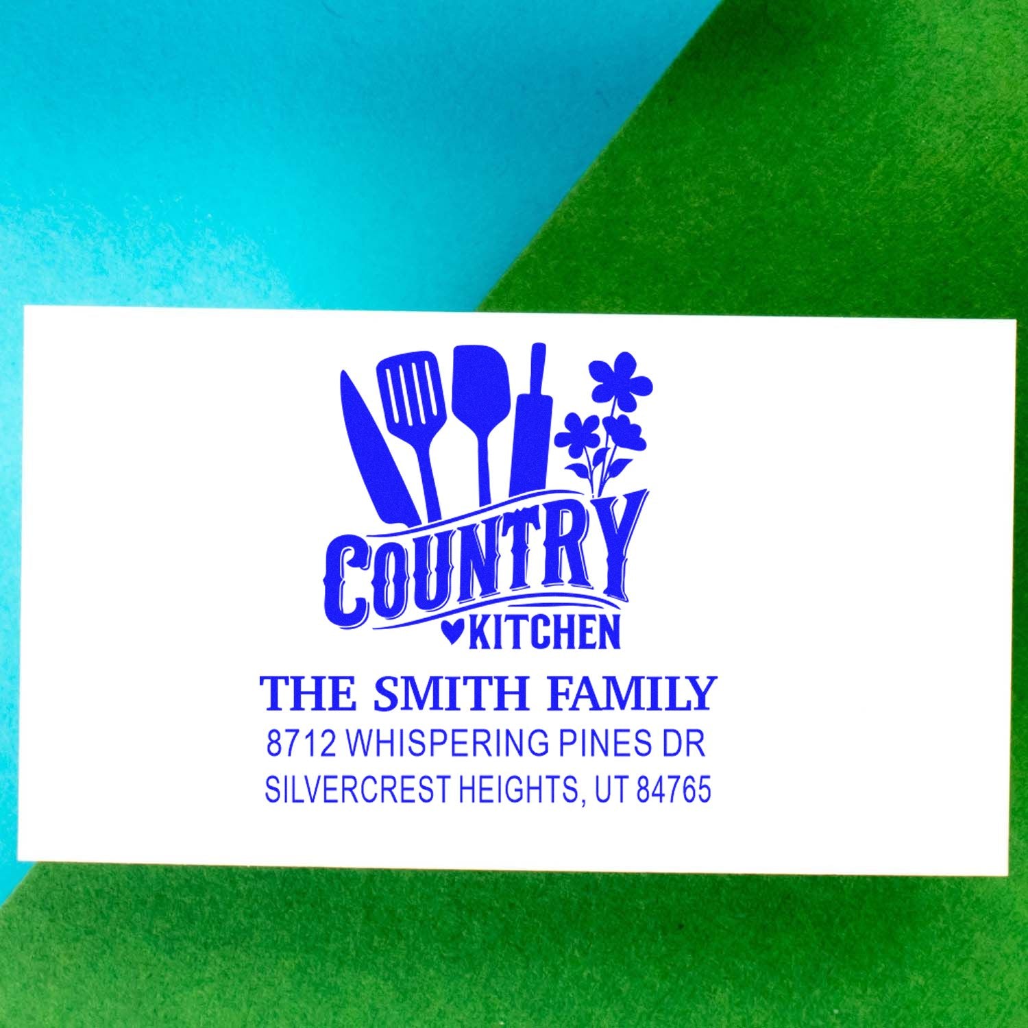 Blue custom country kitchen address stamp with utensils and flowers for The Smith Family, shown on a white card over blue and green background.