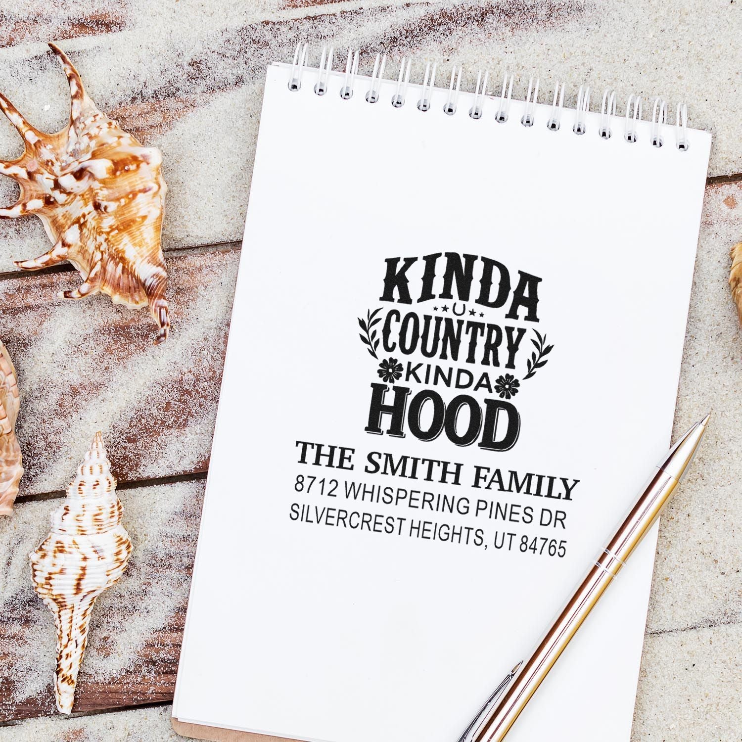 Custom rubber address stamp with Kinda Country Kinda Hood on spiral notepad, styled with seashells and pen on rustic beachwood backdrop.