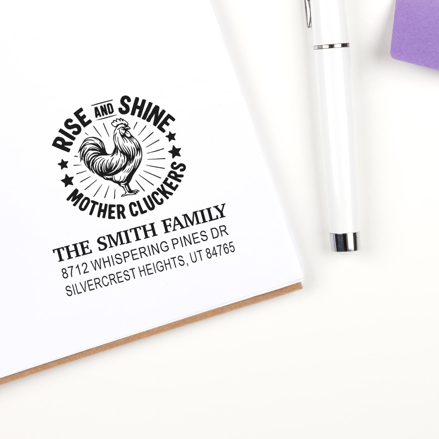 Custom self-inking stamp with rooster and Rise and Shine Mother Cluckers on paper, beside a white pen and notebook.
