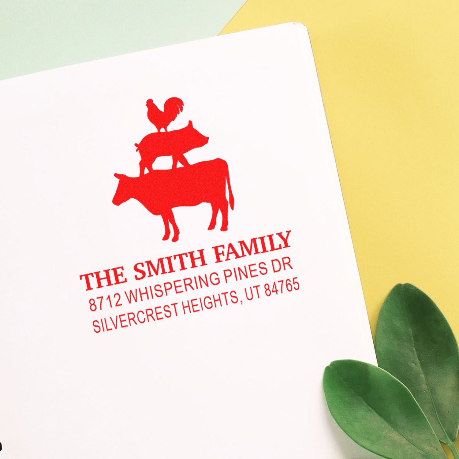 Personalized red family address stamp with pig, cow, and rooster silhouette on white paper, on yellow background with green leaves.
