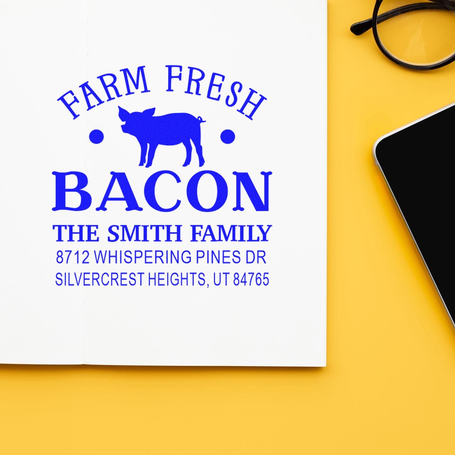 Custom Farm Fresh Bacon address stamp with pig logo on white paper, shown on a yellow desk beside eyeglasses and a smartphone.