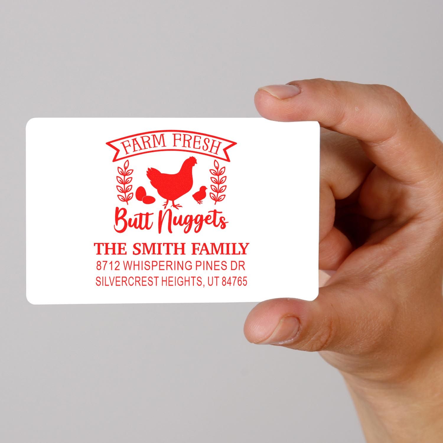 Hand holding custom red farm fresh chicken address stamp with hen graphic, Butt Nuggets text, and The Smith Family address on white card.