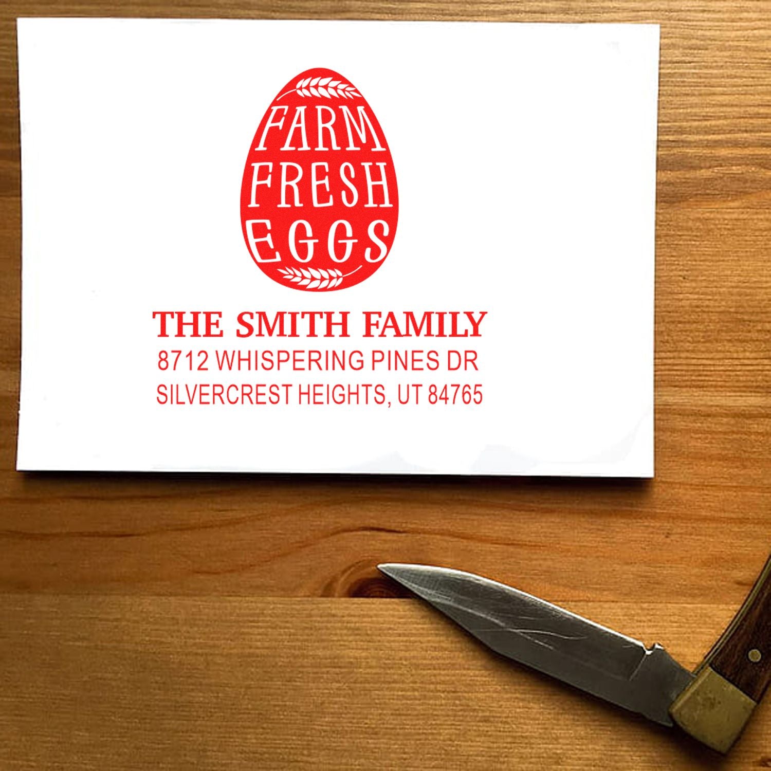 Custom farm fresh eggs address stamp with red egg logo and The Smith Family text on a white card, displayed on a wooden table beside a pocket knife.