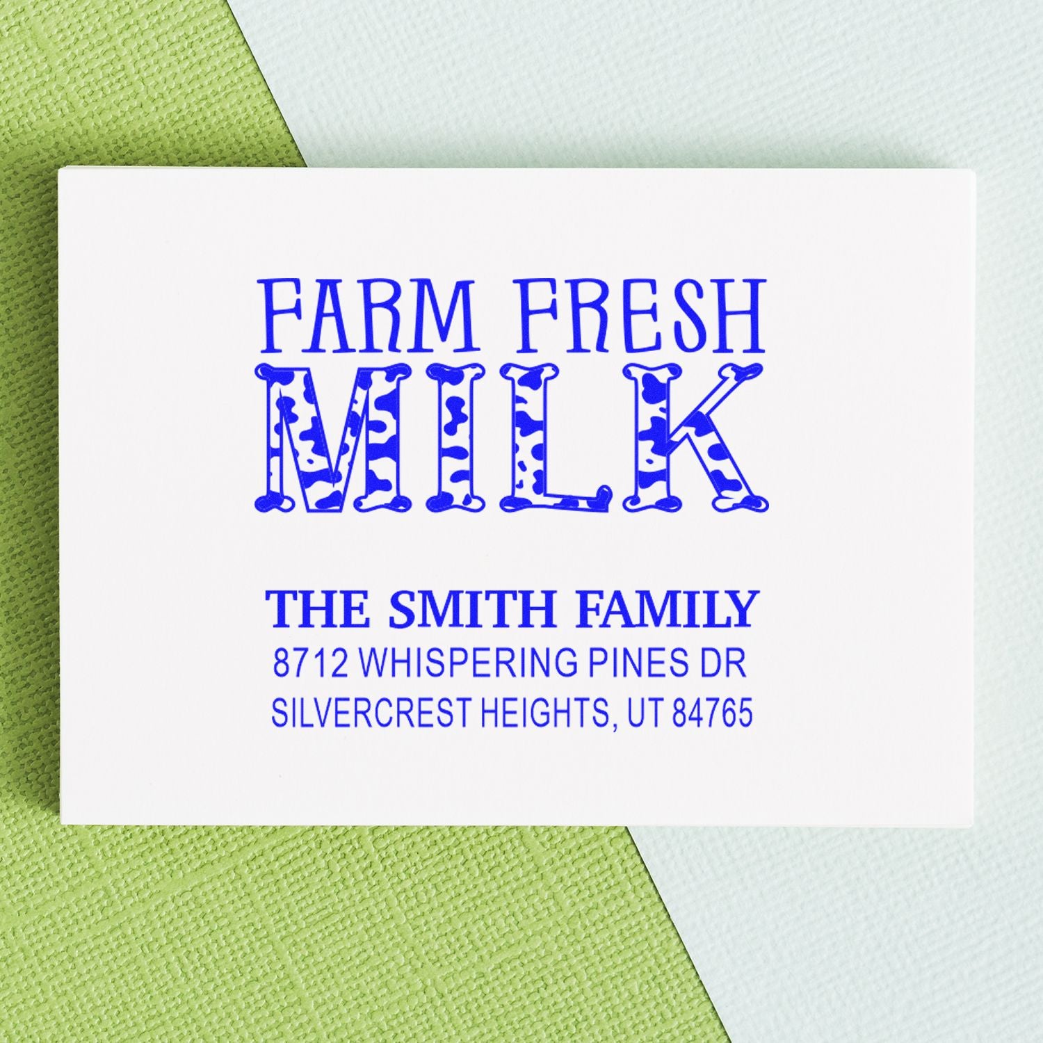 Custom Farm Fresh Milk address stamp with blue cow-print text for The Smith Family, on white card over green and white fabric.