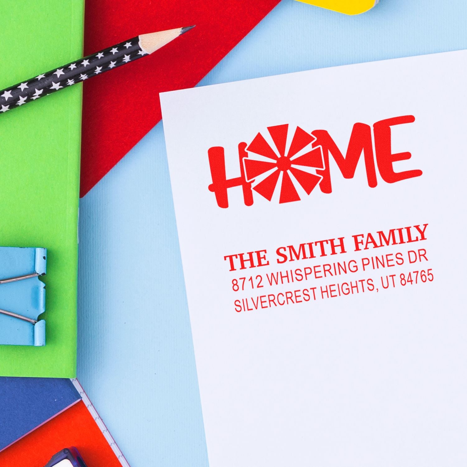 Custom address stamp with HOME design on white paper, surrounded by colorful stationery and a star-pattern pencil on a bright desk.