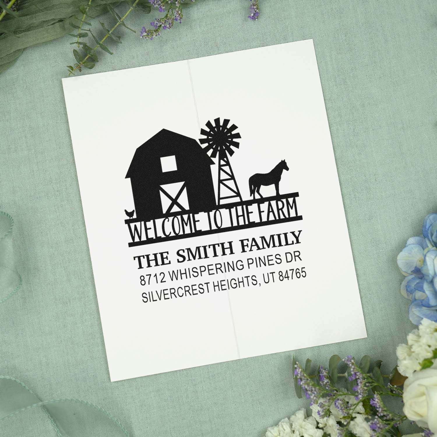 Custom farm welcome address stamp with barn, windmill, horse, and chicken on folded white card, displayed on soft green fabric with flowers.