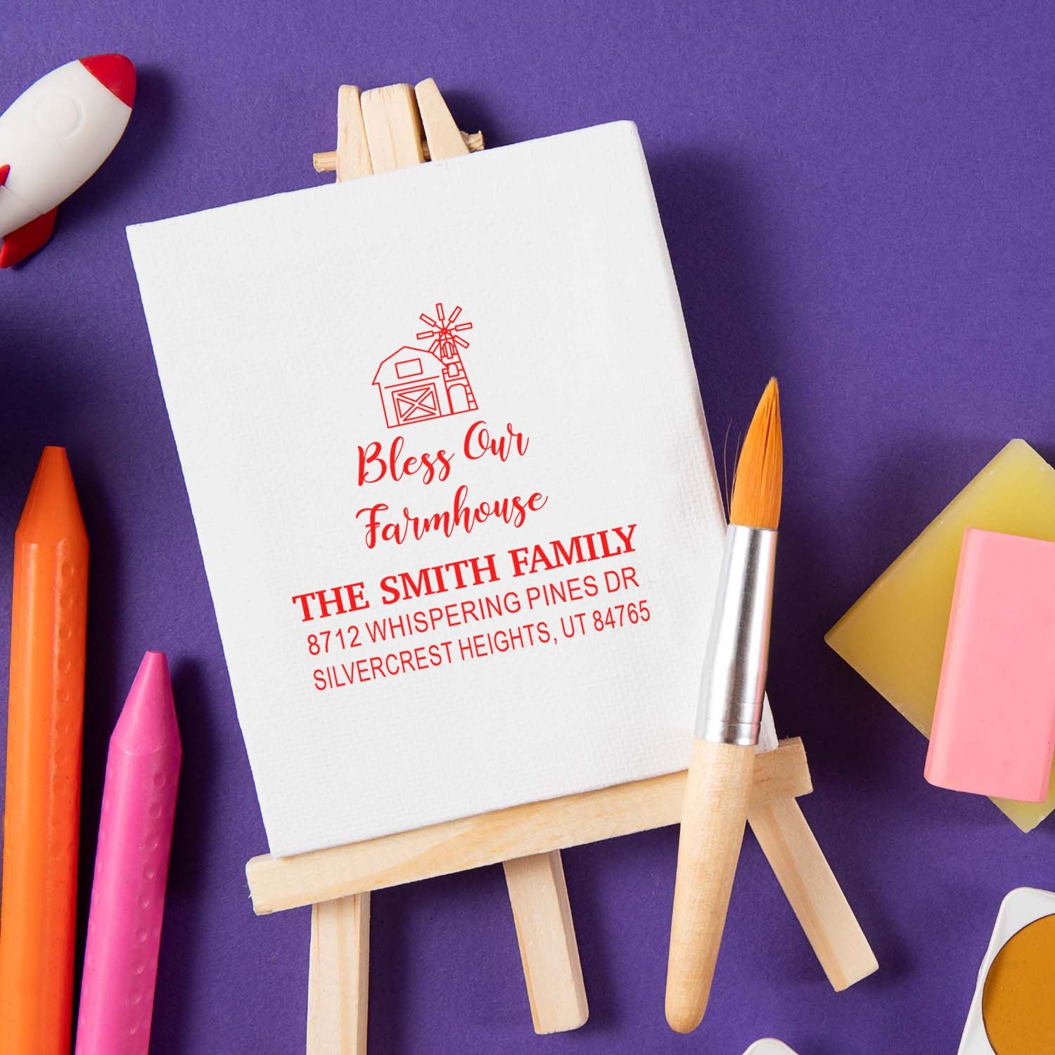 Custom farmhouse address stamp on a wooden easel, with barn icon and family name, surrounded by art supplies on a purple background.