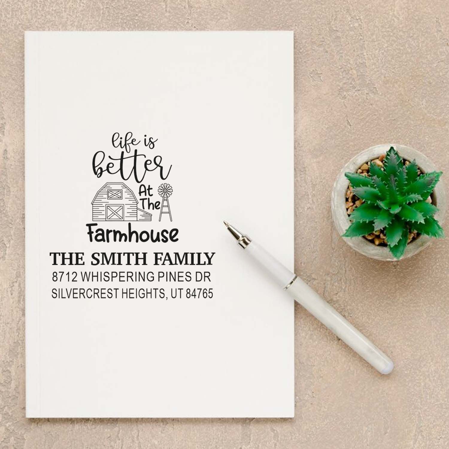 Custom farmhouse return address stamp on white paper with pen, featuring Smith family name, barn design, and address beside a small succulent.