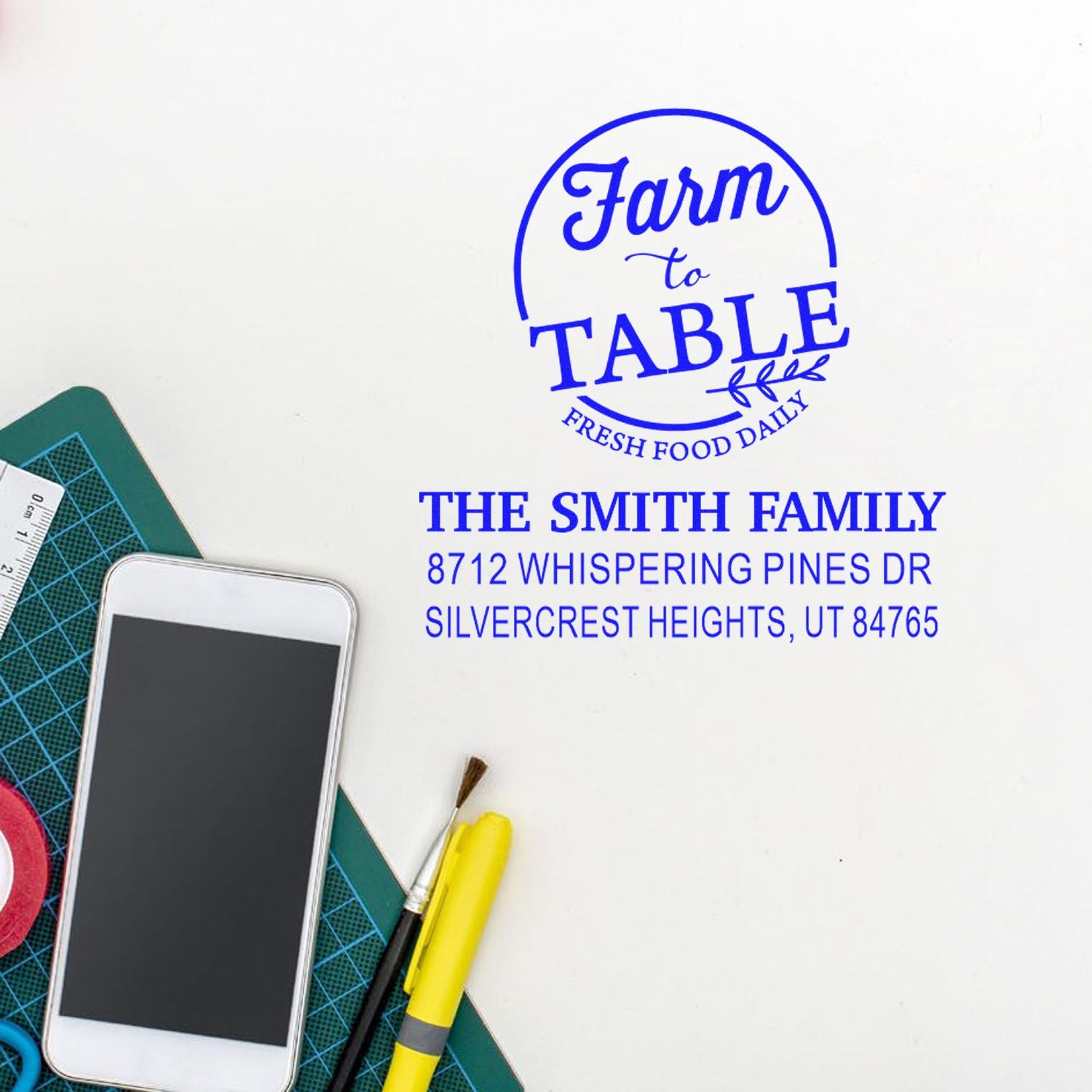 Custom blue farm-to-table return address stamp on white paper, beside craft tools, ruler, and smartphone on a workspace.
