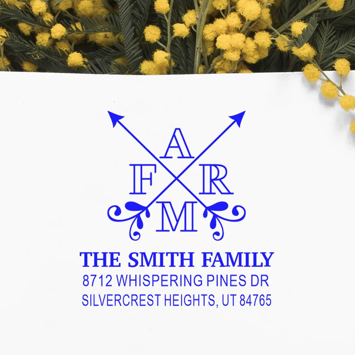 Custom blue monogram address stamp with crossed arrows, The Smith Family name, and home address on a white card with yellow mimosa flowers.