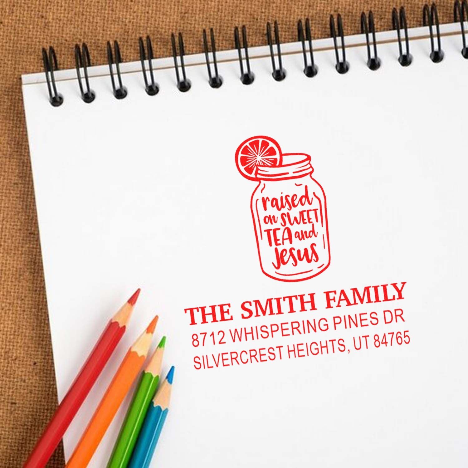 Personalized self-inking stamp with mason jar raised on sweet tea and Jesus design on a spiral notebook, beside colorful pencils.