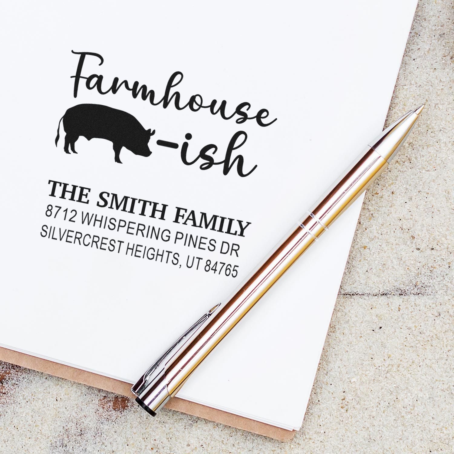 Custom farmhouse-style address stamp with pig graphic and Farmhouse-ish text on paper, shown with a gold pen on a notebook.