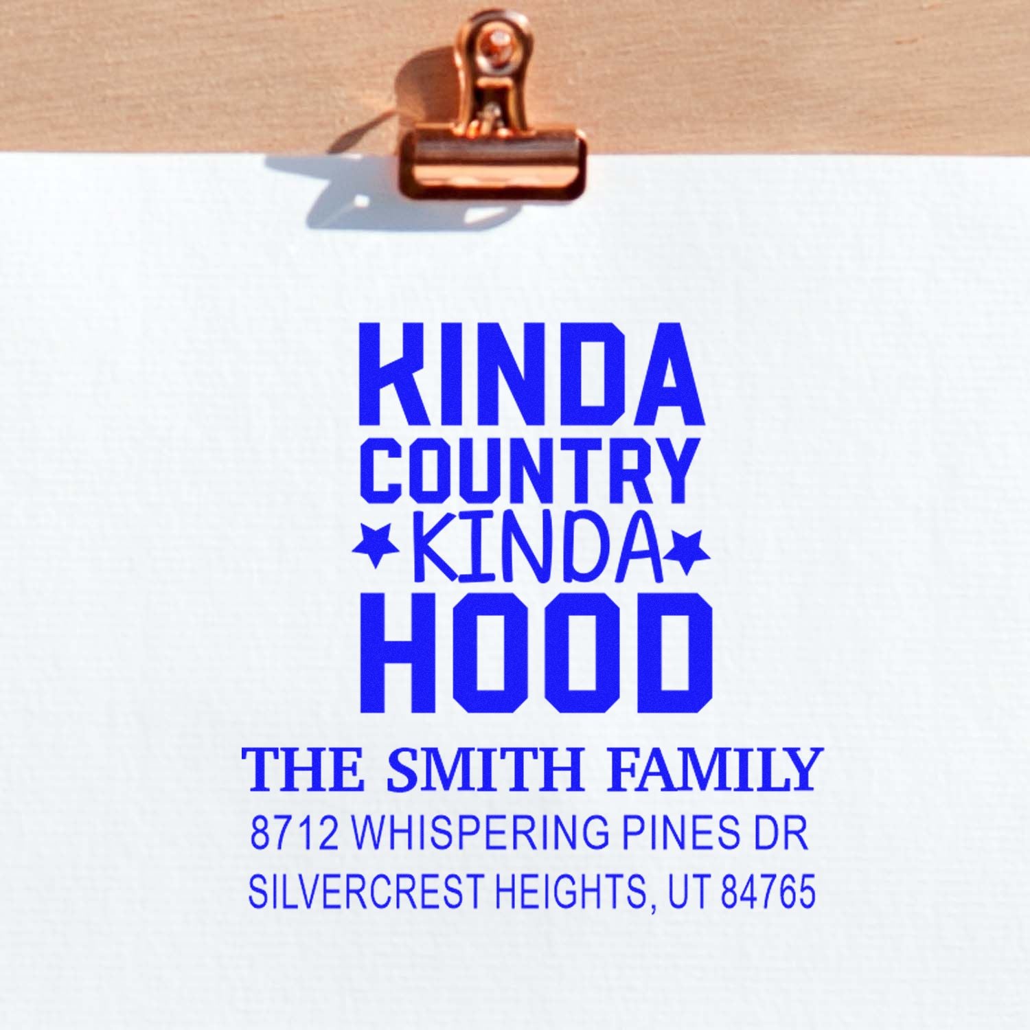 Custom rubber stamp on white paper clipped to clipboard, reading Kinda Country Kinda Hood with The Smith Family address in blue.