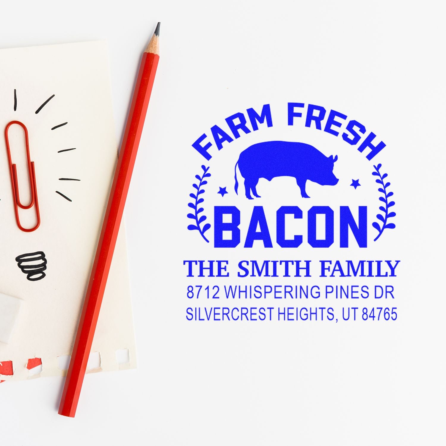 Custom farm fresh bacon return address stamp with pig graphic, family name, and address beside a red pencil and paper clips.