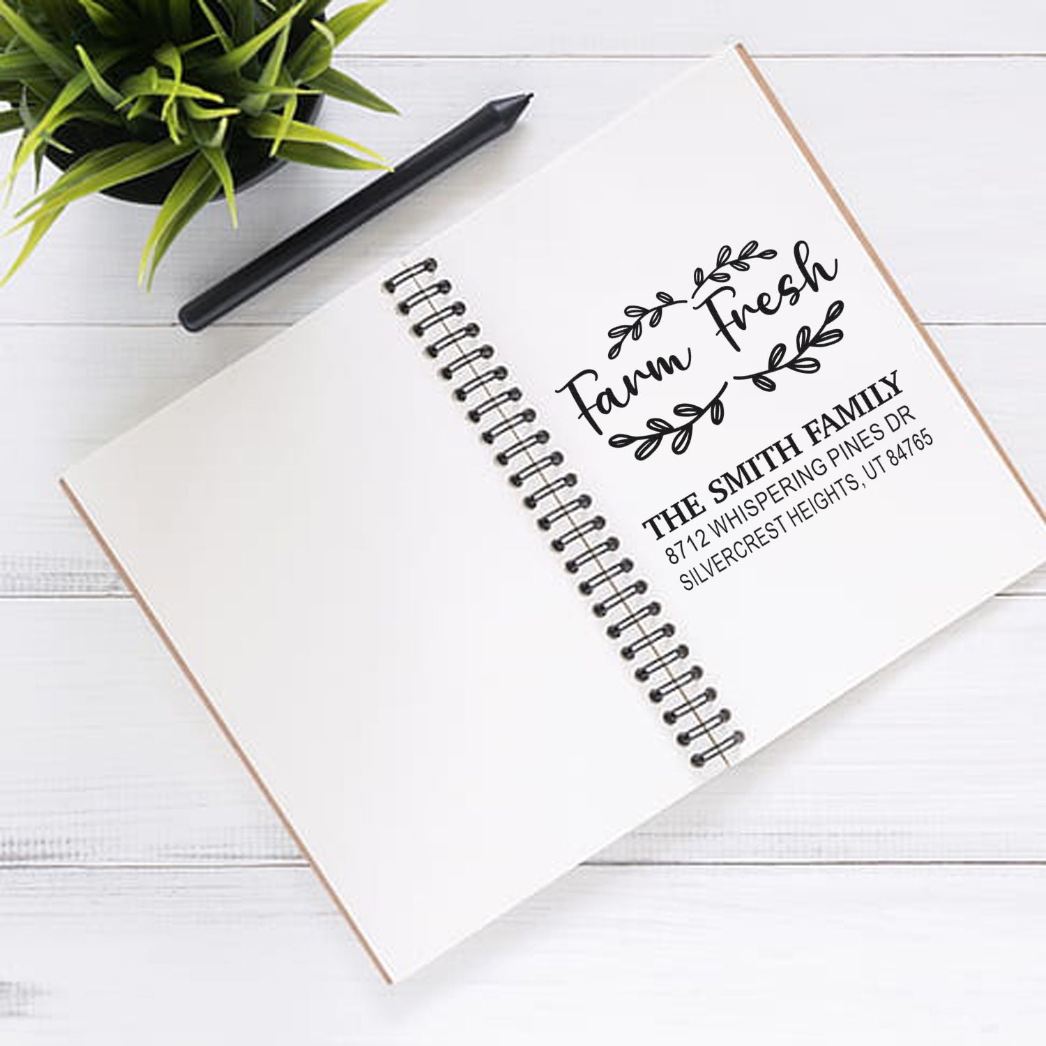 Custom family address stamp on a spiral notebook on a white desk, beside a pen and potted plant, featuring Farm Fresh and The Smith Family return address.
