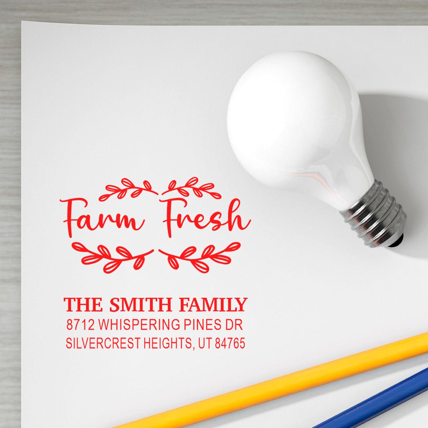Custom return address stamp mockup with Farm Fresh design and Smith family address on white paper, shown beside a light bulb and pencils.