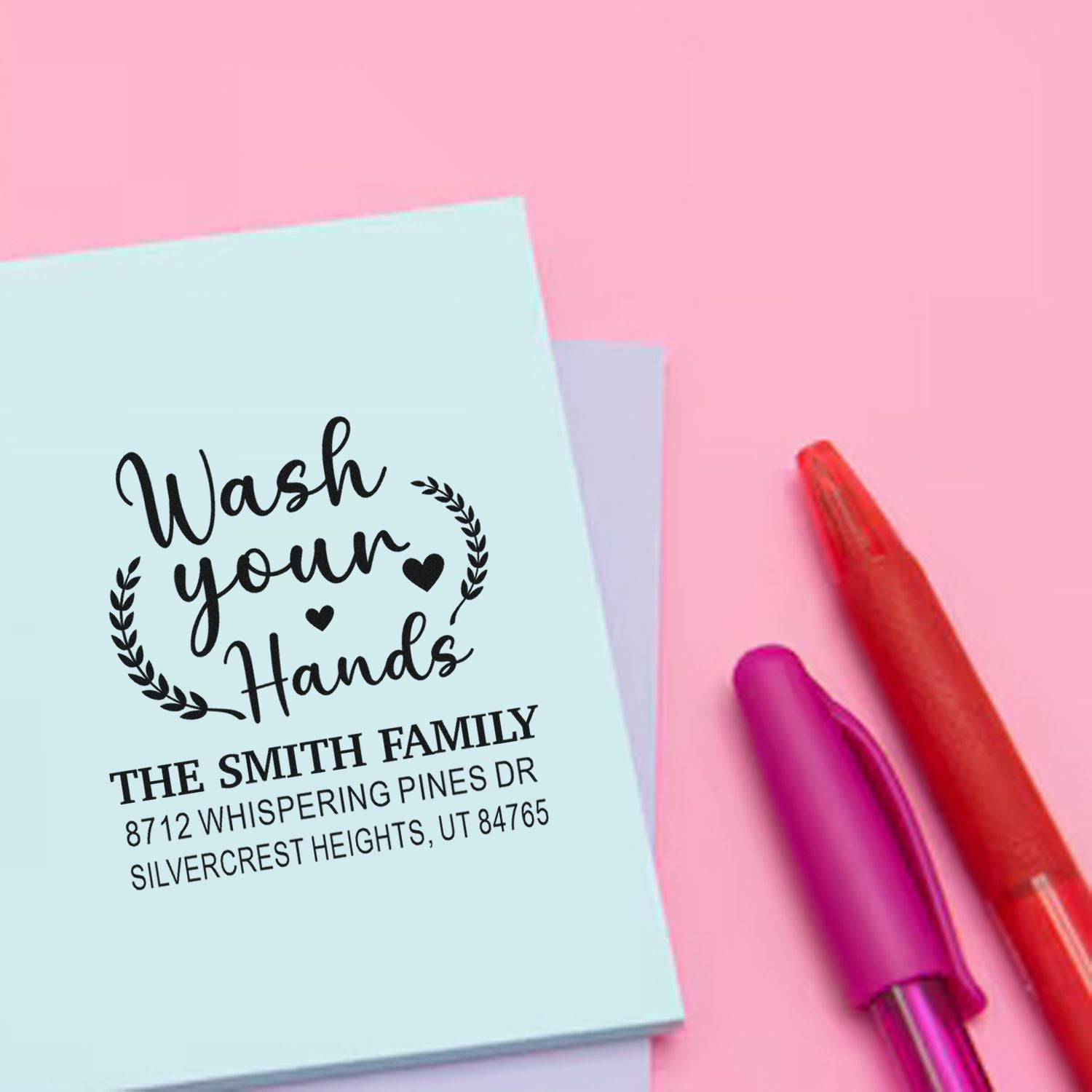 Custom address stamp with Wash your Hands design on pastel paper, beside colorful pens on a pink background.