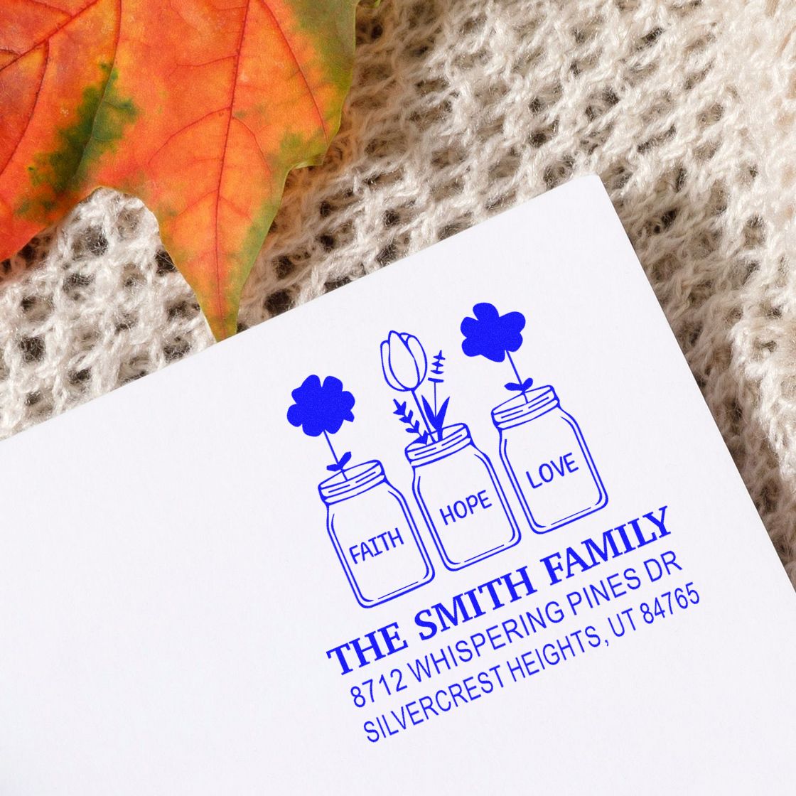 Blue custom family address stamp with faith, hope, love mason jars and flowers on white paper, styled on a knitted fabric background with autumn leaf.
