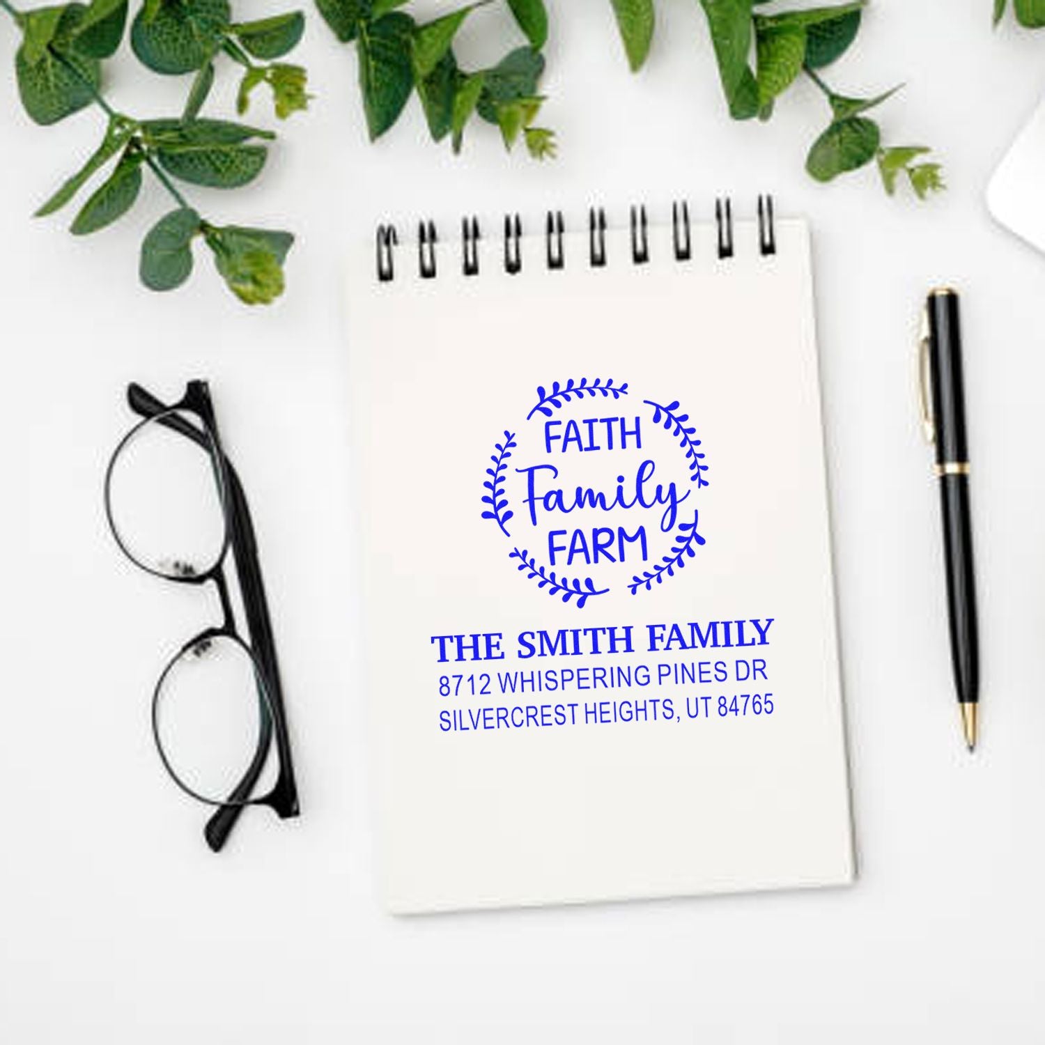 Custom Faith Family Farm return address stamp on spiral notebook, with glasses, pen and greenery on a white desk.
