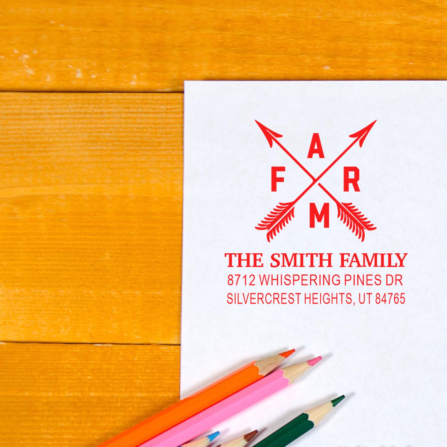 Custom family address stamp on white paper with crossed arrows logo, resting on a wooden table beside colorful pencils.