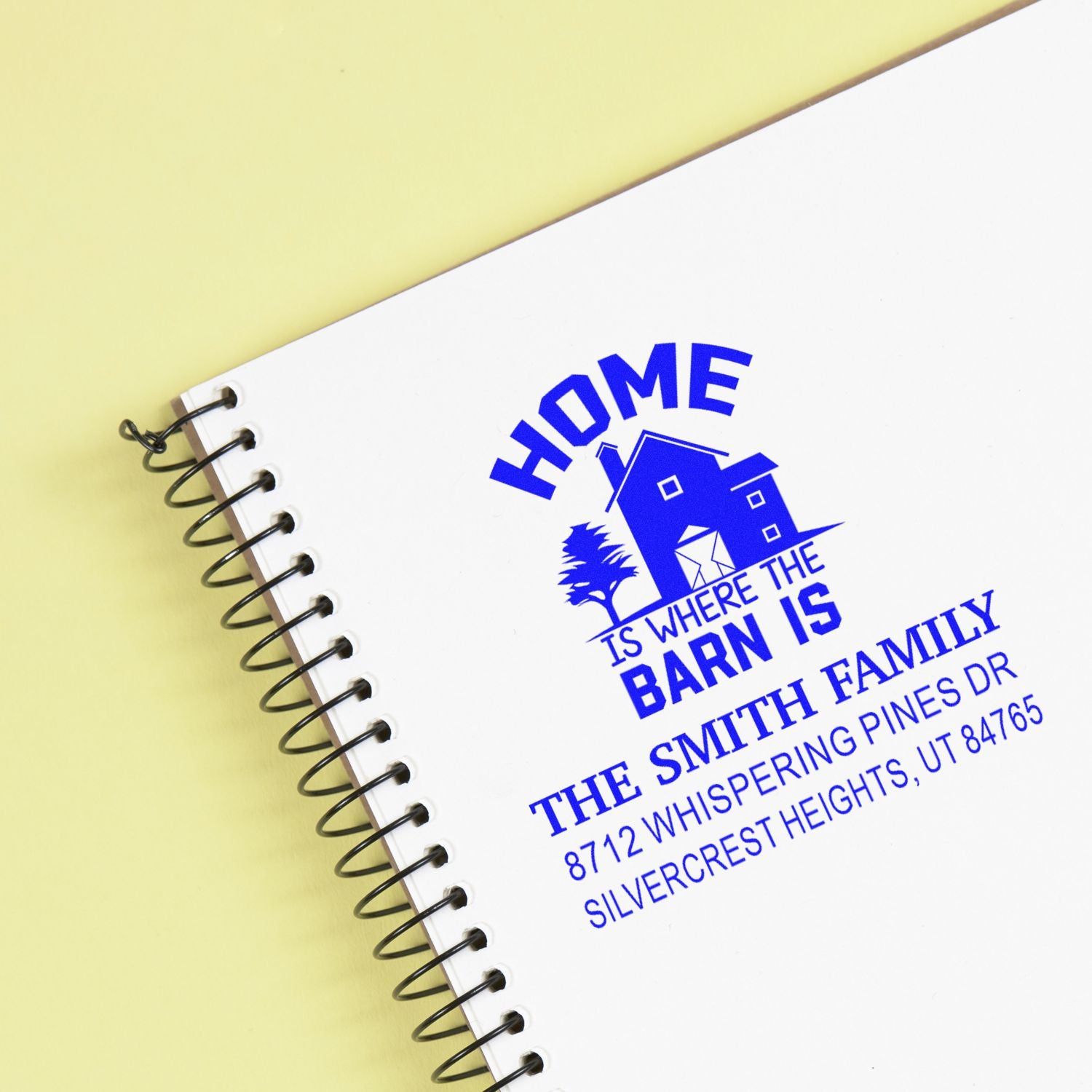Personalized blue address stamp on a spiral-bound notebook, featuring Home Is Where the Barn Is with house and barn graphic on a pale yellow background.