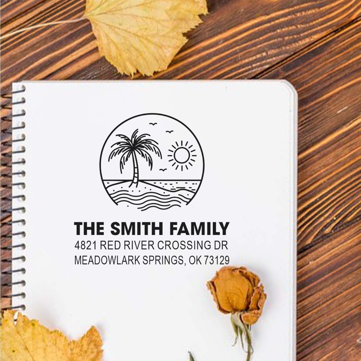 Personalized family address stamp with palm tree, sun and beach scene on a notebook, styled on a wooden table with autumn leaves.