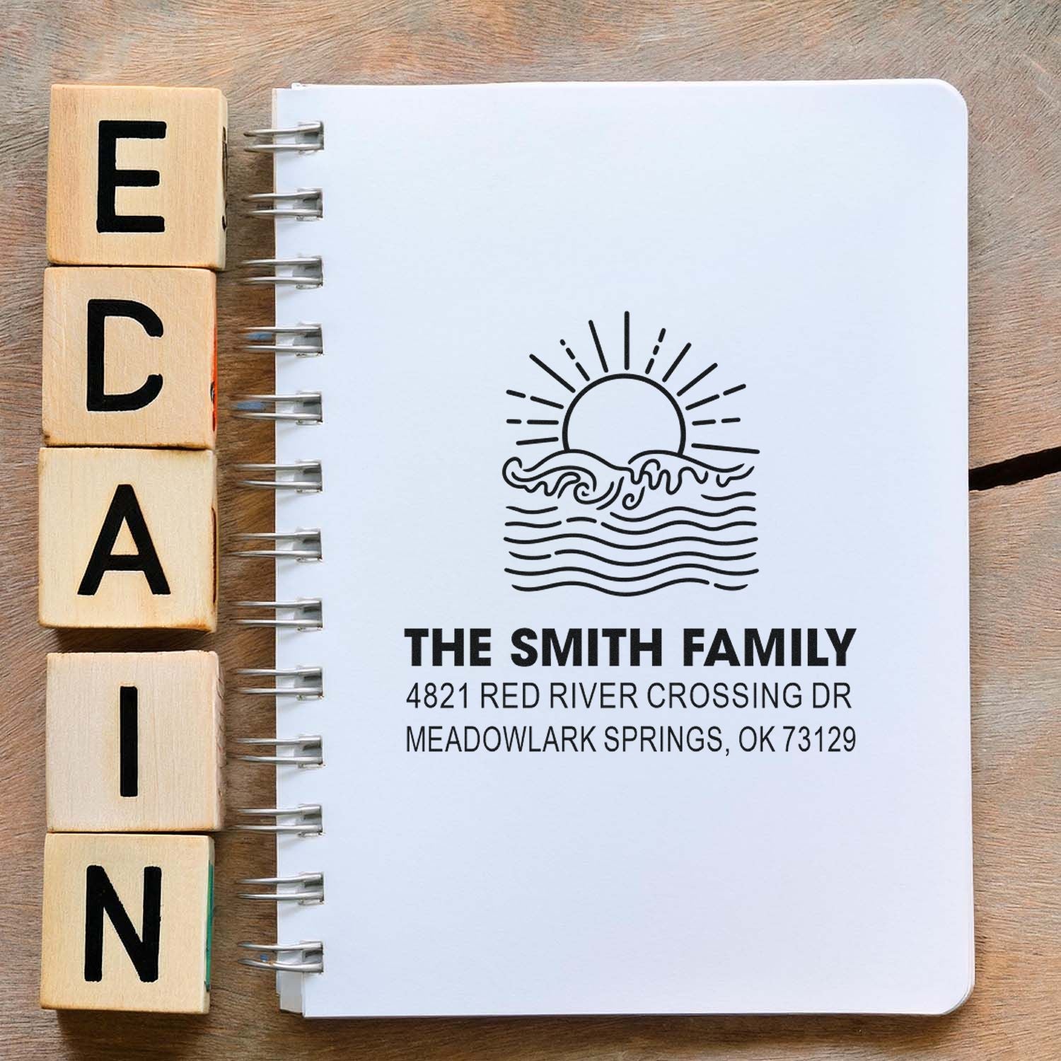 Custom family address stamp on a spiral notebook beside wooden letter stamps, featuring a sun-and-waves logo and The Smith Family address.