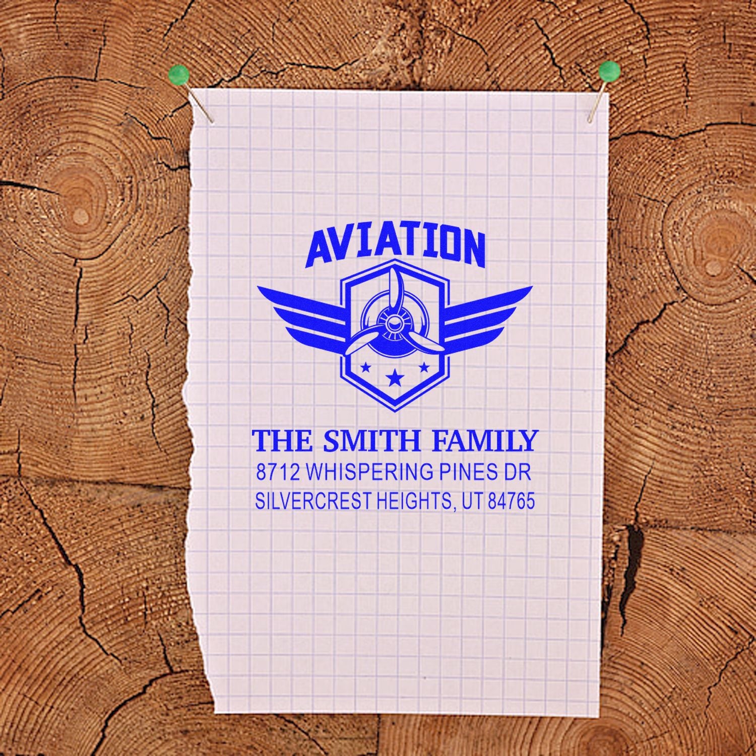 Custom aviation return address stamp with blue winged propeller design on graph paper, pinned to a rustic wood background.