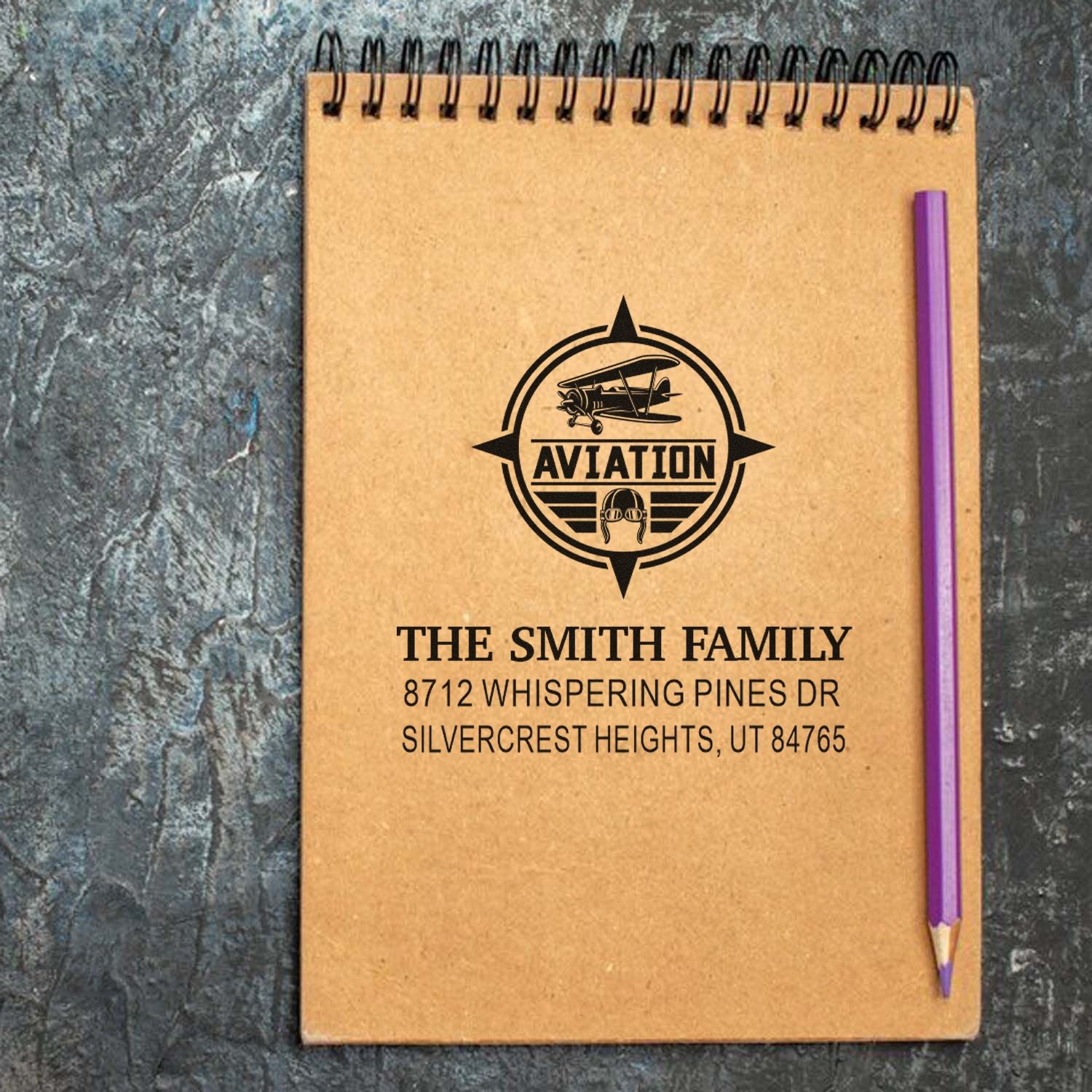 Personalized aviation address stamp on a kraft spiral notebook, featuring a biplane logo, family name, address, and a purple pencil.