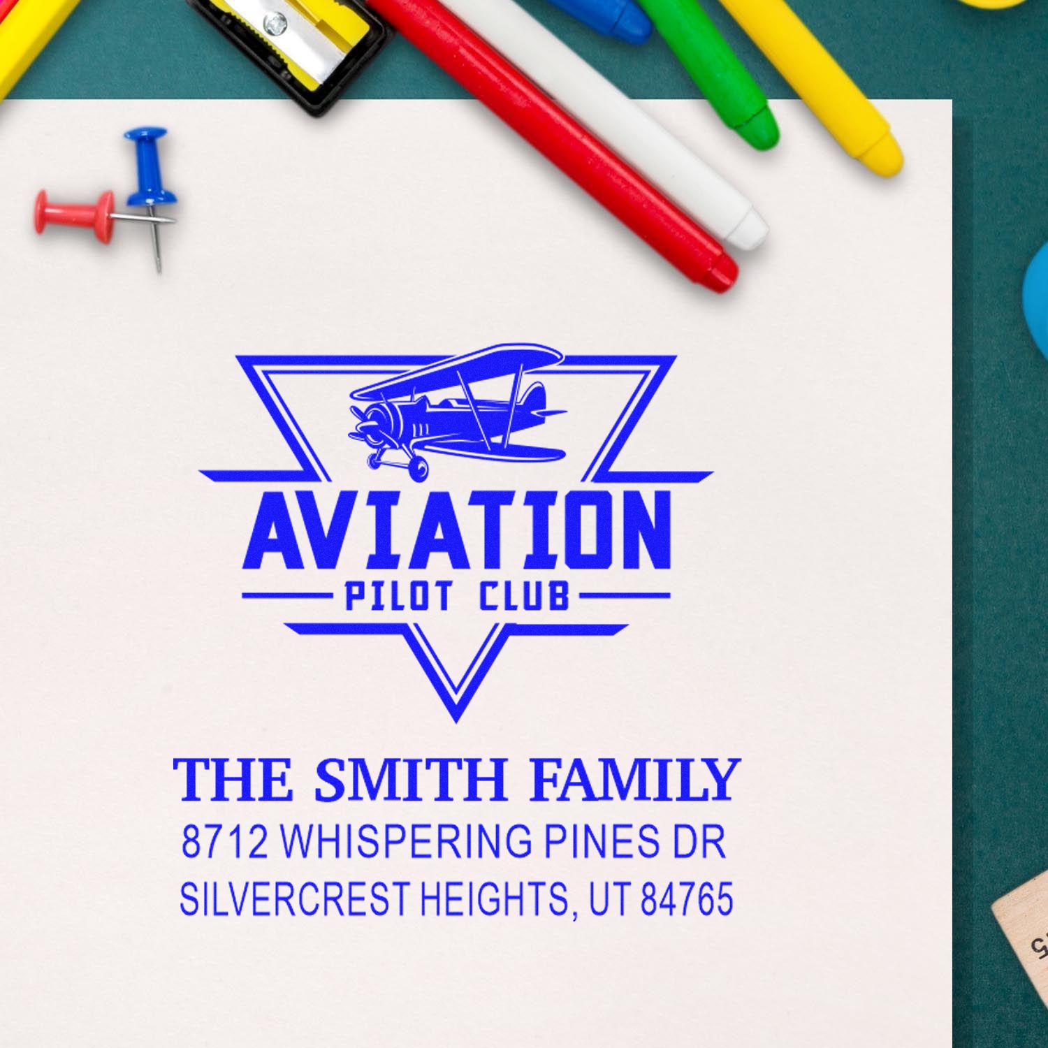 Custom aviation pilot club self-inking stamp with biplane logo, The Smith Family name and address on white paper, surrounded by colorful pens and push pins.