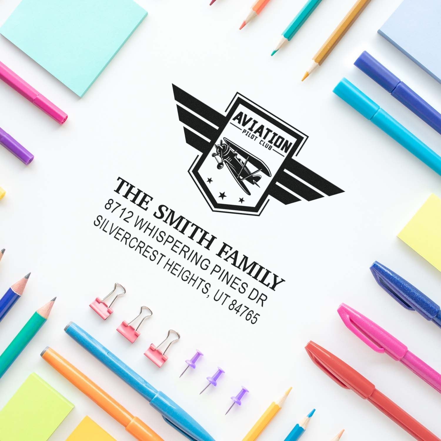 Custom aviation pilot club stamp on white paper, surrounded by colorful pens, pencils, sticky notes and binder clips.