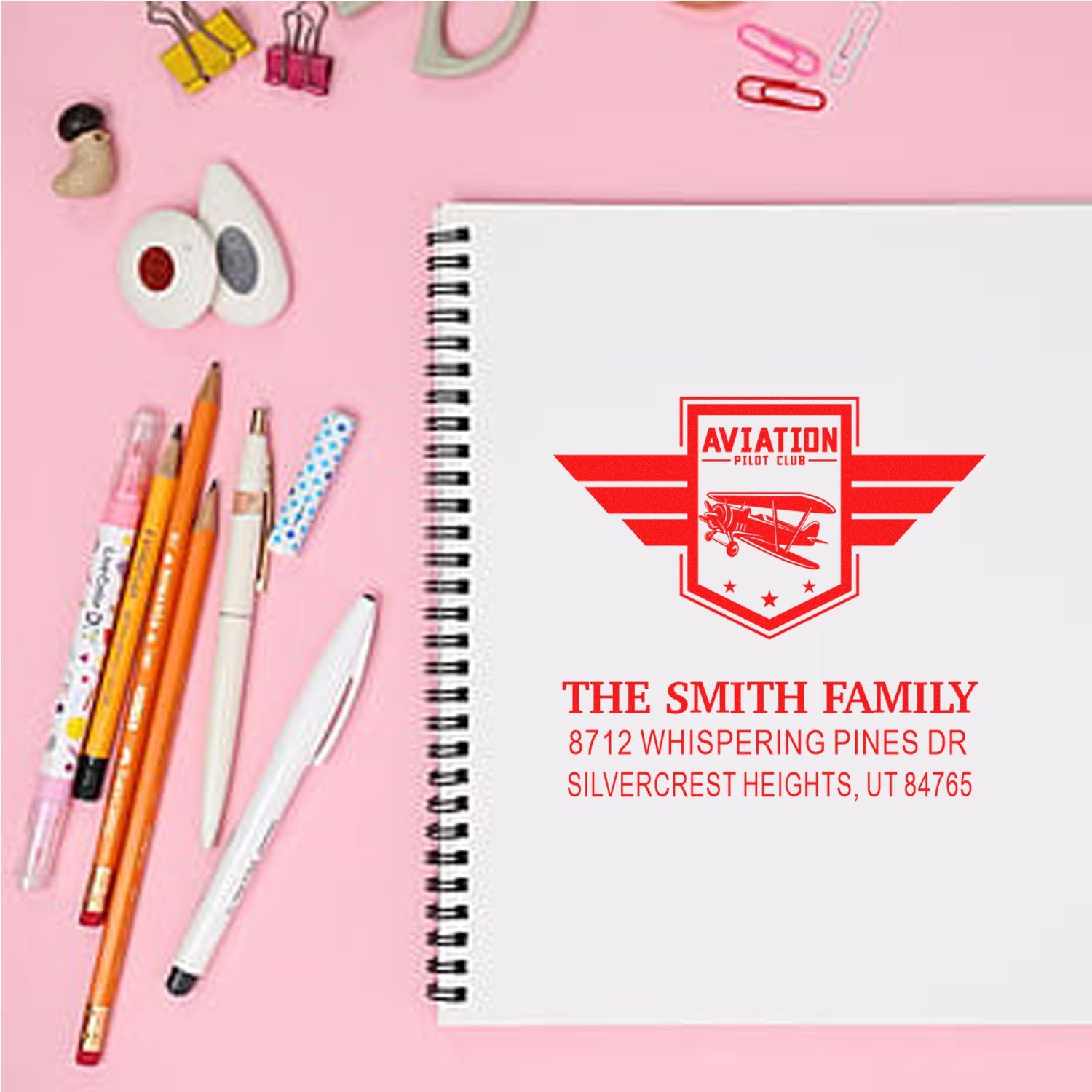 Aviation Pilot Club personalized address stamp on a white spiral notebook, surrounded by pencils, pens, and stationery on a pink desk.