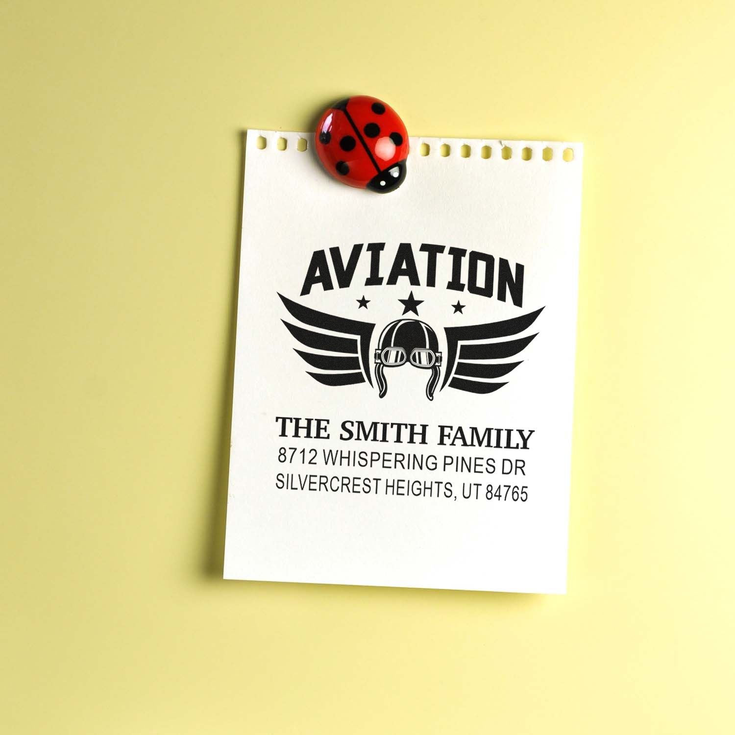 Aviation-themed family address stamp on white notepaper, clipped with a ladybug magnet against a pale yellow background.