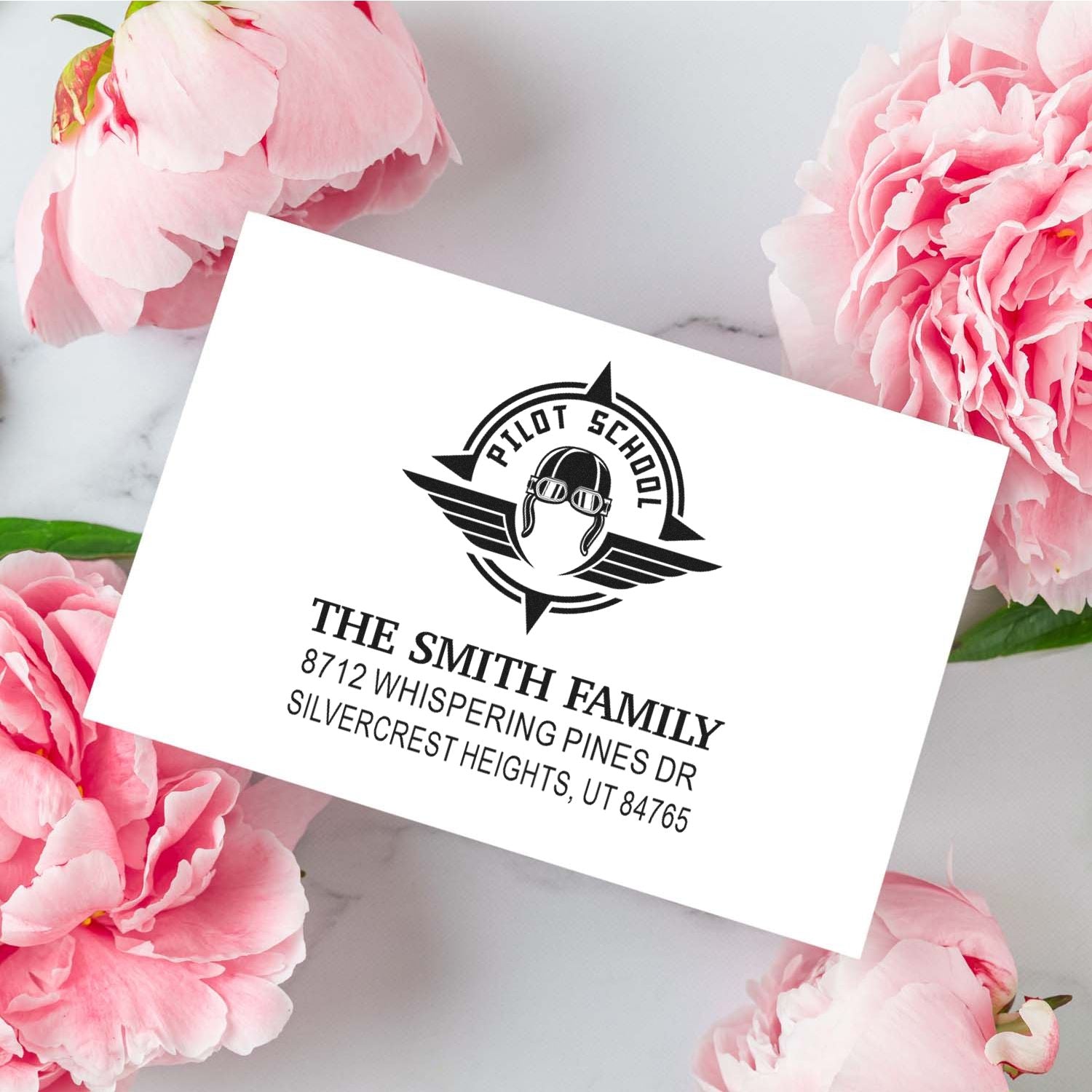 Custom return address stamp for The Smith Family with Pilot School logo, shown on a white card among pink peonies on a marble background.