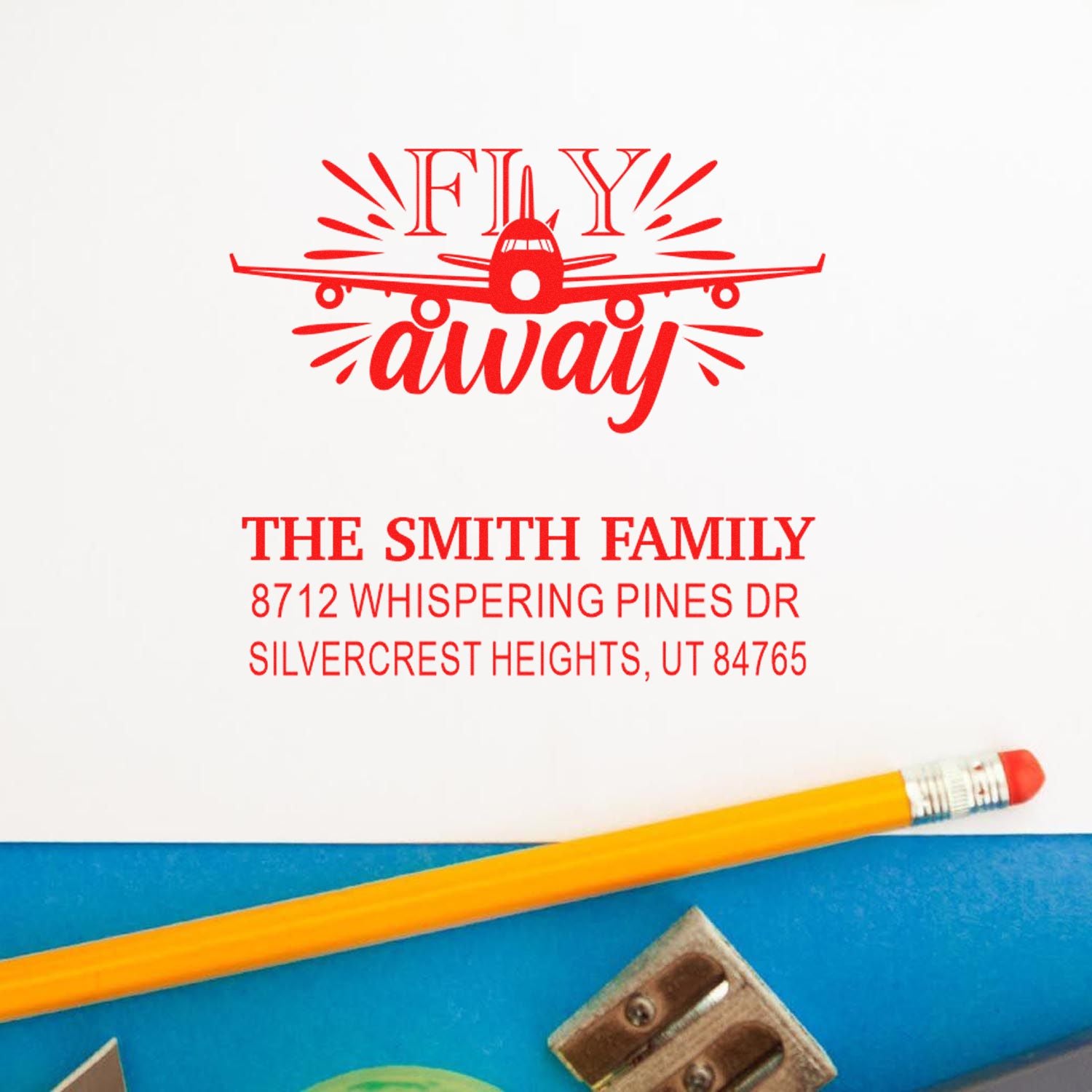 Custom red airplane mail-away address stamp with Fly away for The Smith Family, displayed on white paper beside a pencil.