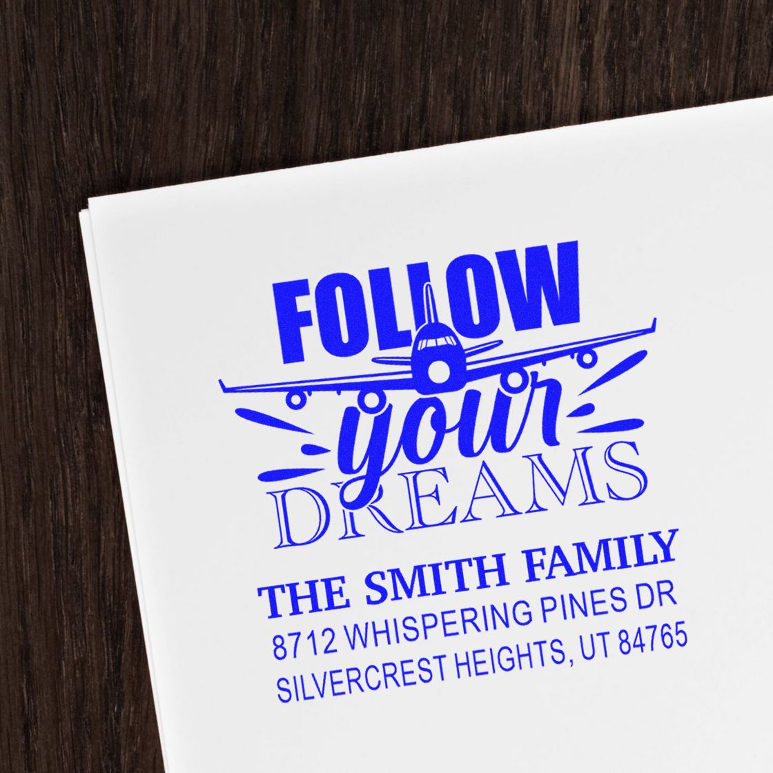 Custom blue self-inking stamp on white paper with airplane graphic, Follow Your Dreams, and Smith family address on a wood tabletop.